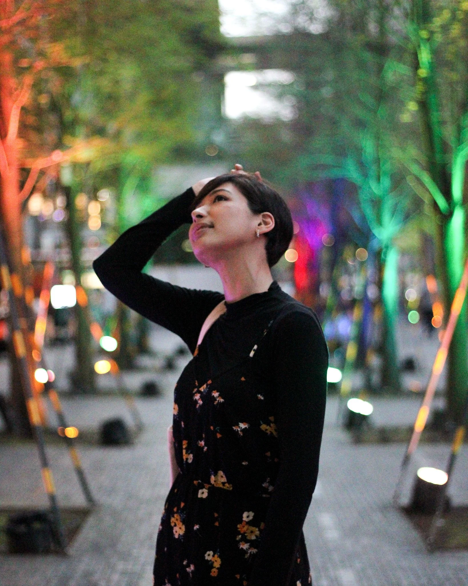 A woman with short dark hair and earrings stands outdoors at night, with colorful lights illuminating trees in the background, touching her head and looking upward.