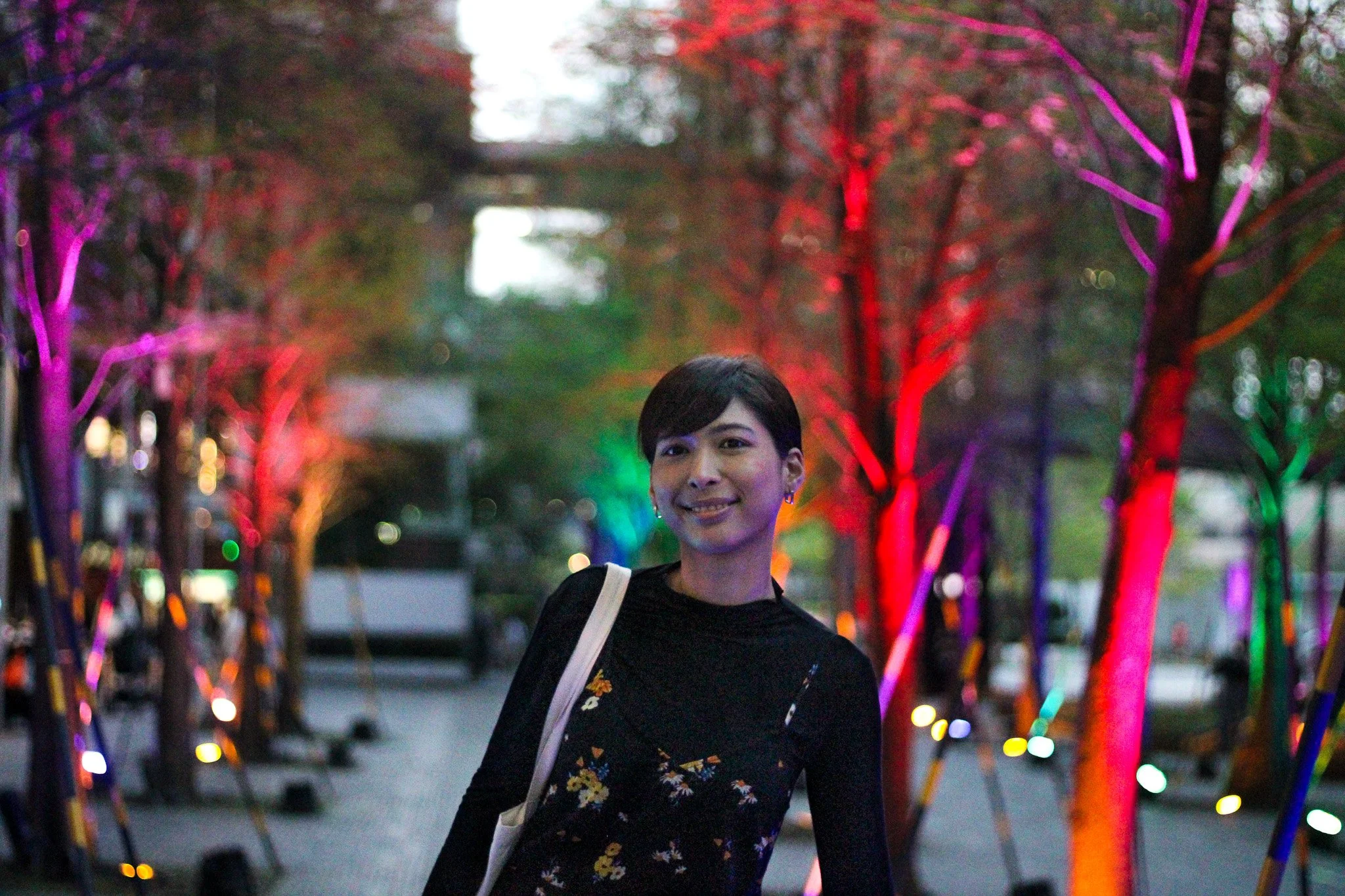 A woman with short black hair smiling at night in front of trees lit with colorful purple, red, green, and orange lights.