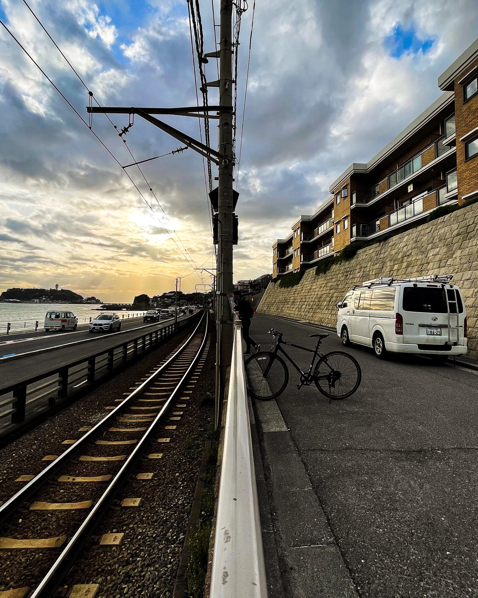 View of a seaside road at sunset with train tracks, cars, a bicycle, and a man standing by the rails. Buildings with balconies are on the right, and the sky is partly cloudy.