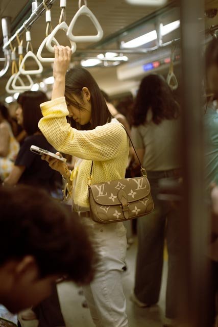 身穿黃色針織衫的女子在擁擠捷運車廂裡拉著吊環滑手機，台北