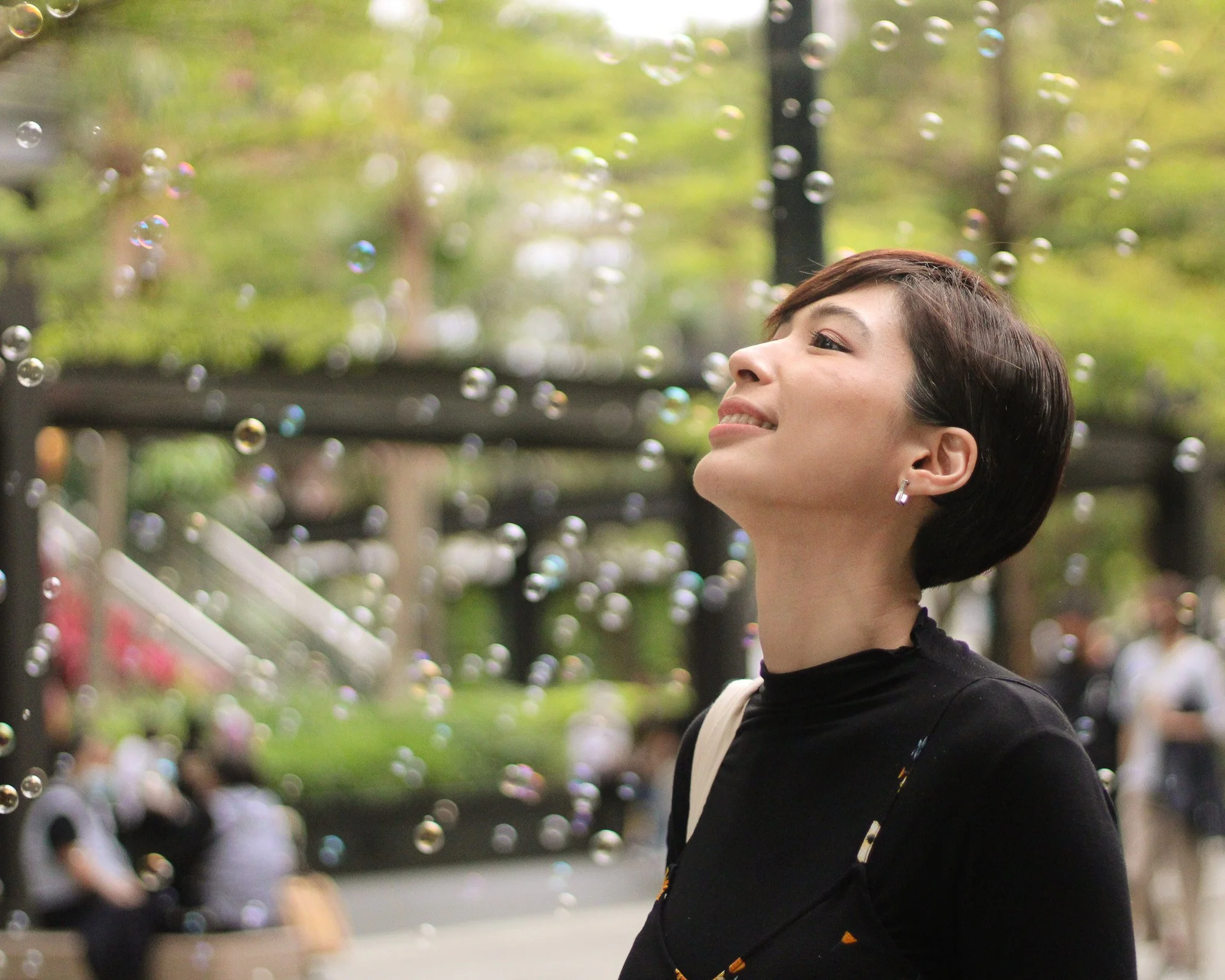 A woman smiling and standing outdoors in a park on a sunny day with bubbles floating around her, blurred people and trees in the background.