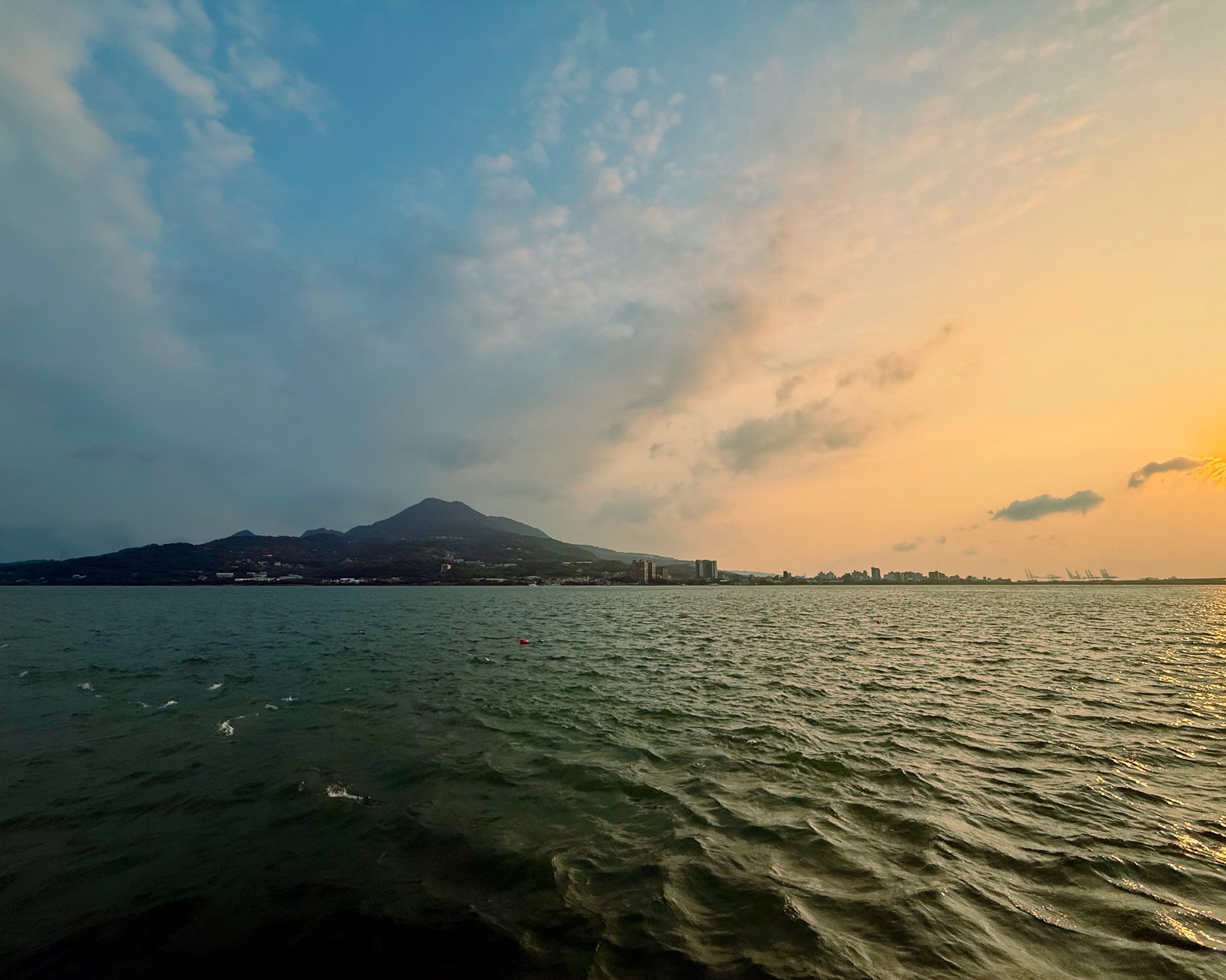夕陽下的河口水面泛起波紋，山巒、岸邊建築與遠方港口起重機橫列天際，淡水
