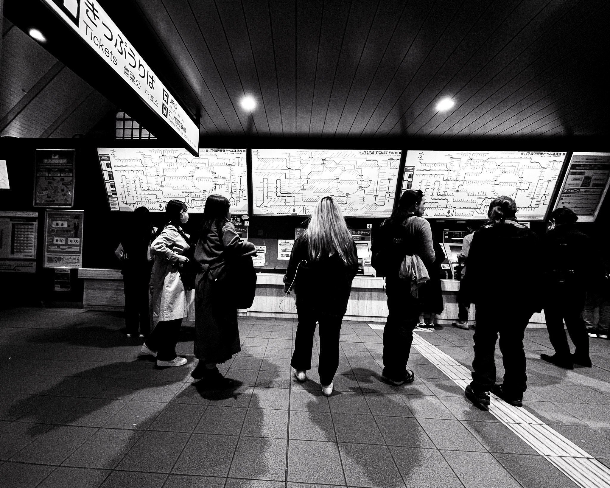 People waiting in line at a train station ticket counter with large maps and signs, some wearing masks.