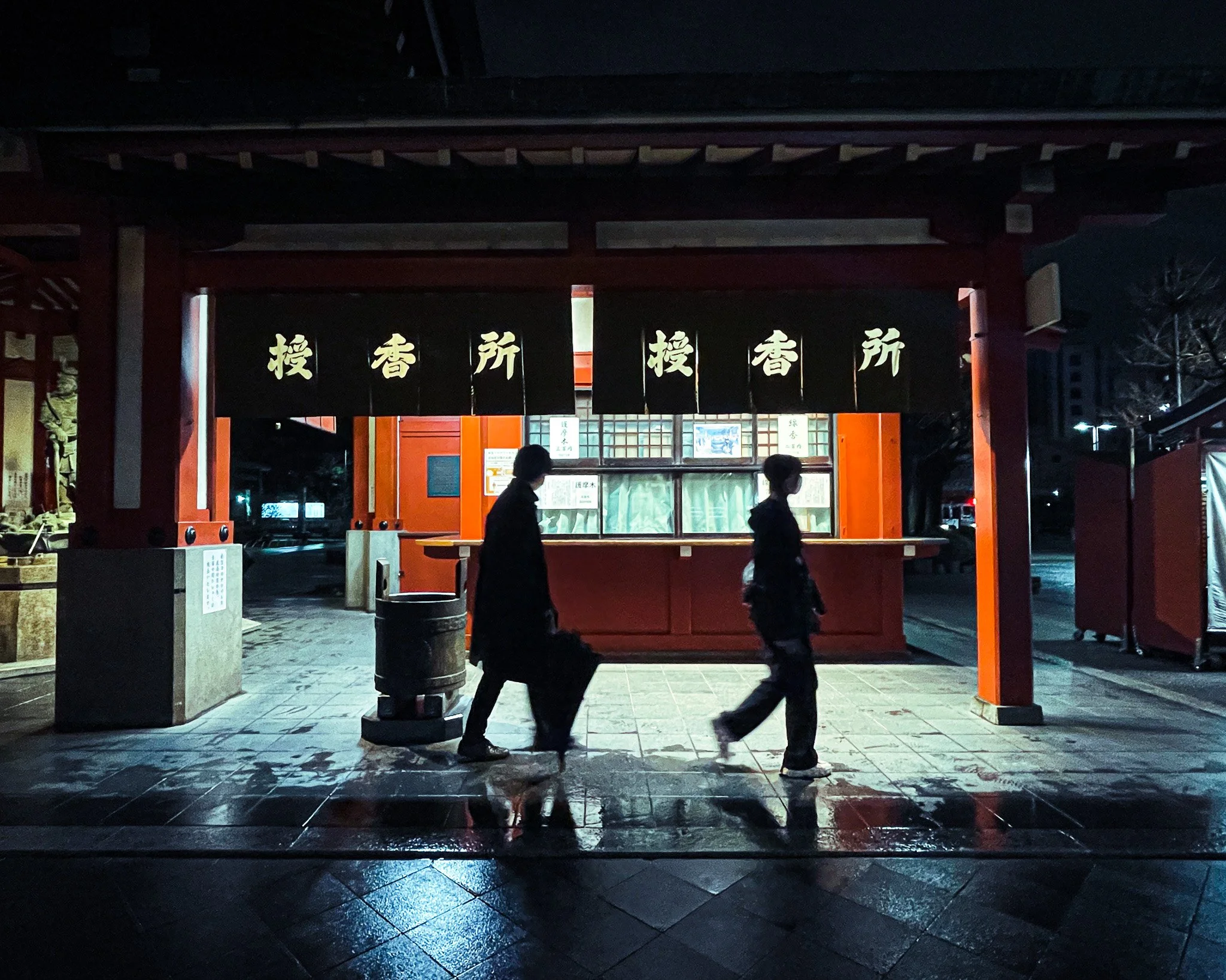 Two silhouetted people walk past a covered food or ticket stand at night, under a Japanese torii gate with black hanging banners with gold characters in Japanese.