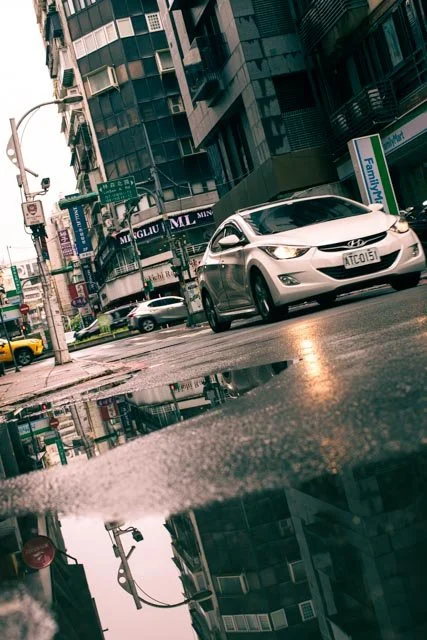 低角度街景中，白色轎車與商店招牌倒映在雨後積水裡，台灣