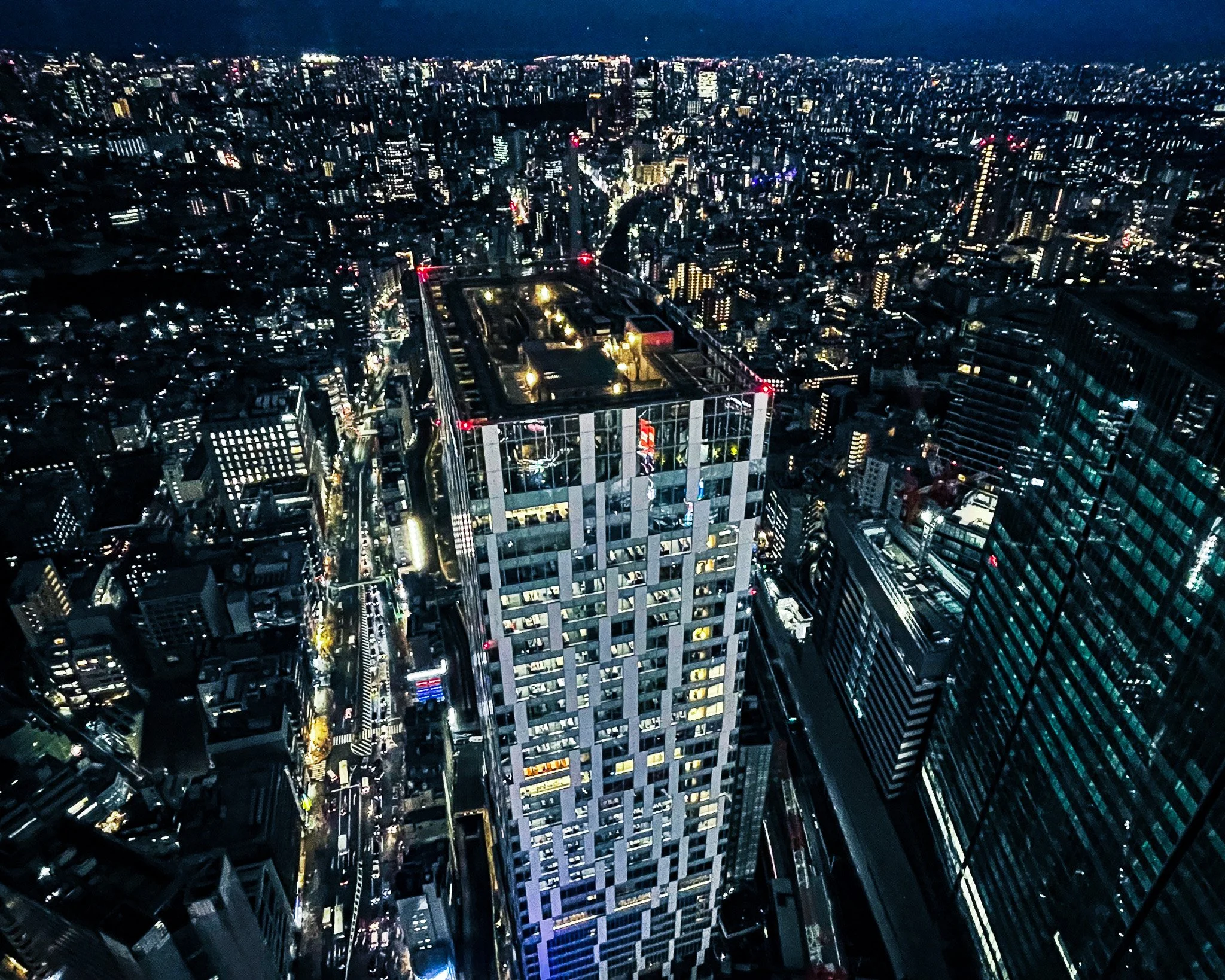 Nighttime aerial view of a cityscape with illuminated skyscrapers and streets, with a prominent high-rise building in the foreground.