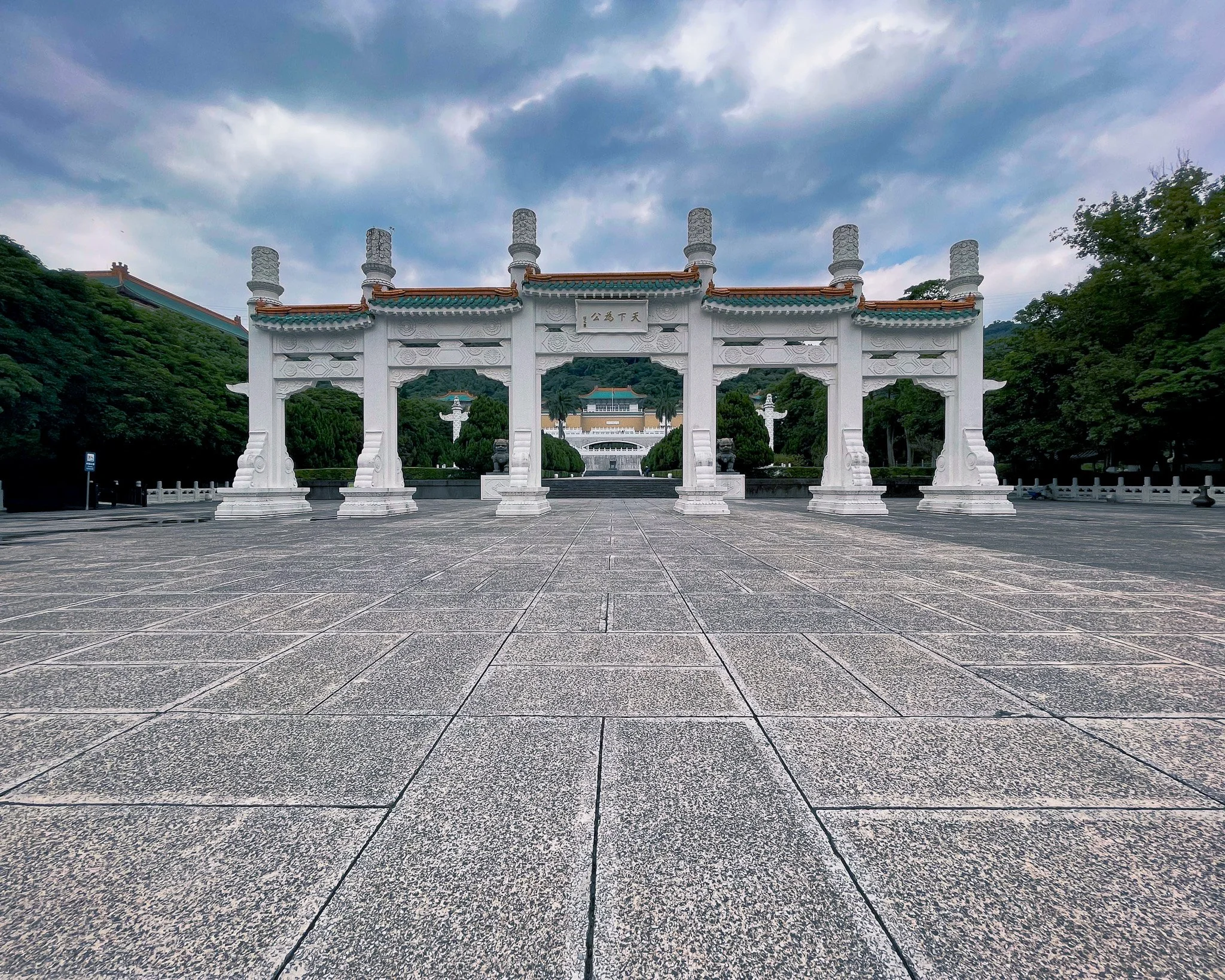 An ornate, white Chinese-style archway with five large supporting pillars, set on a paved stone courtyard. In the background, green trees and a building are visible, with dark clouds overhead.