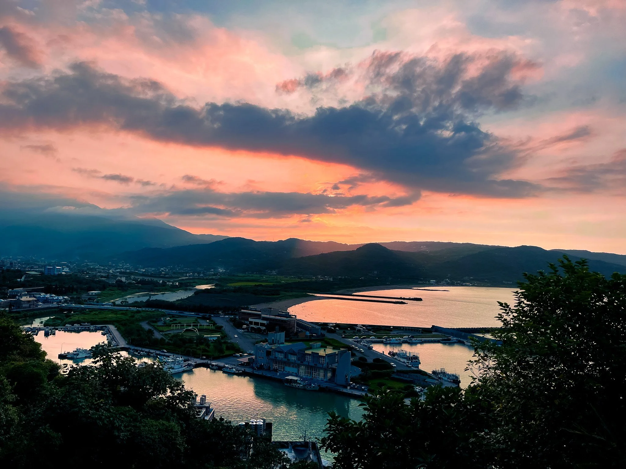 Scenic view of a harbor with boats, surrounded by greenery and hills, under a colorful sunset sky with clouds.