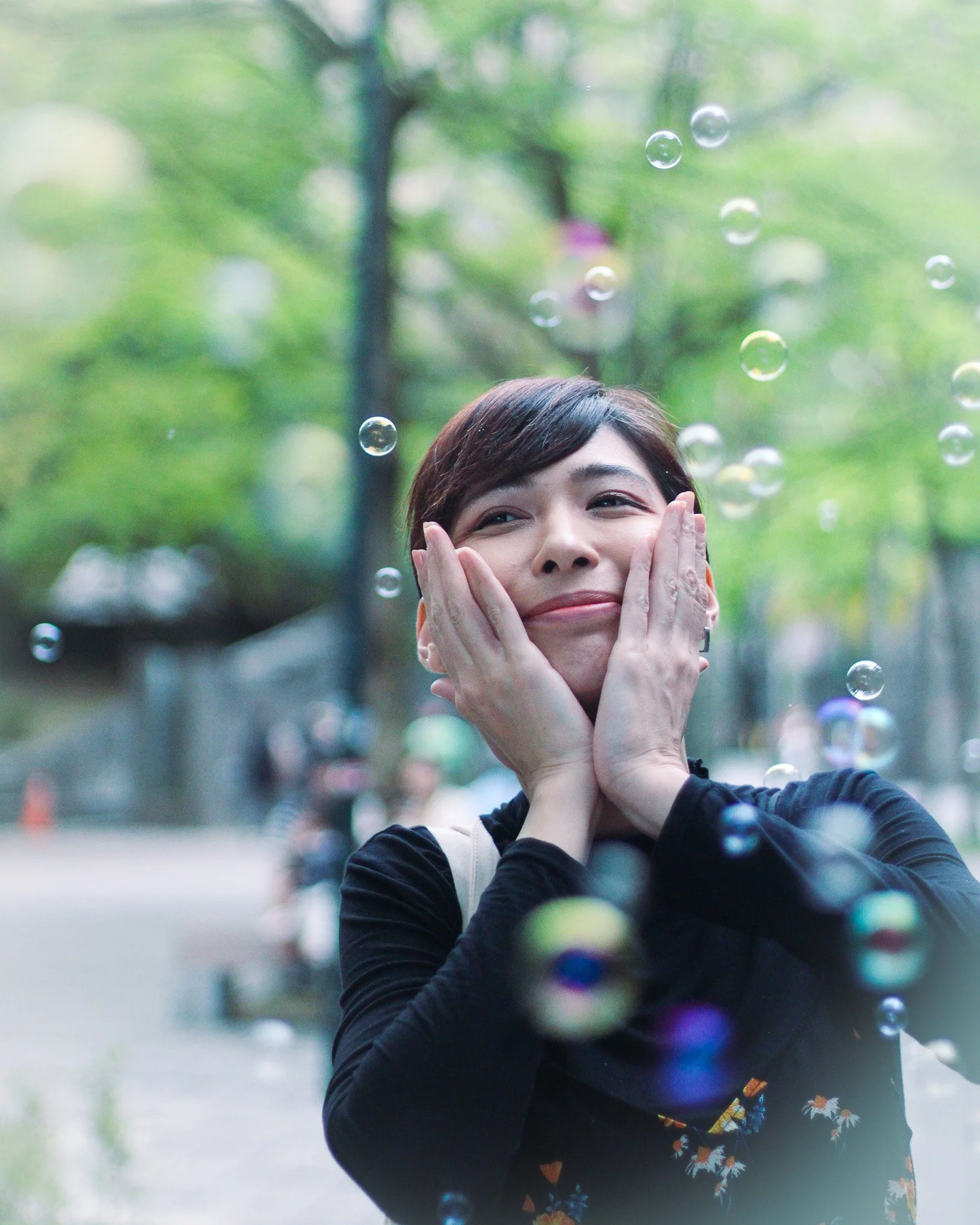 Woman smiling and holding her face with both hands outdoors, surrounded by floating soap bubbles, with green trees in the background.
