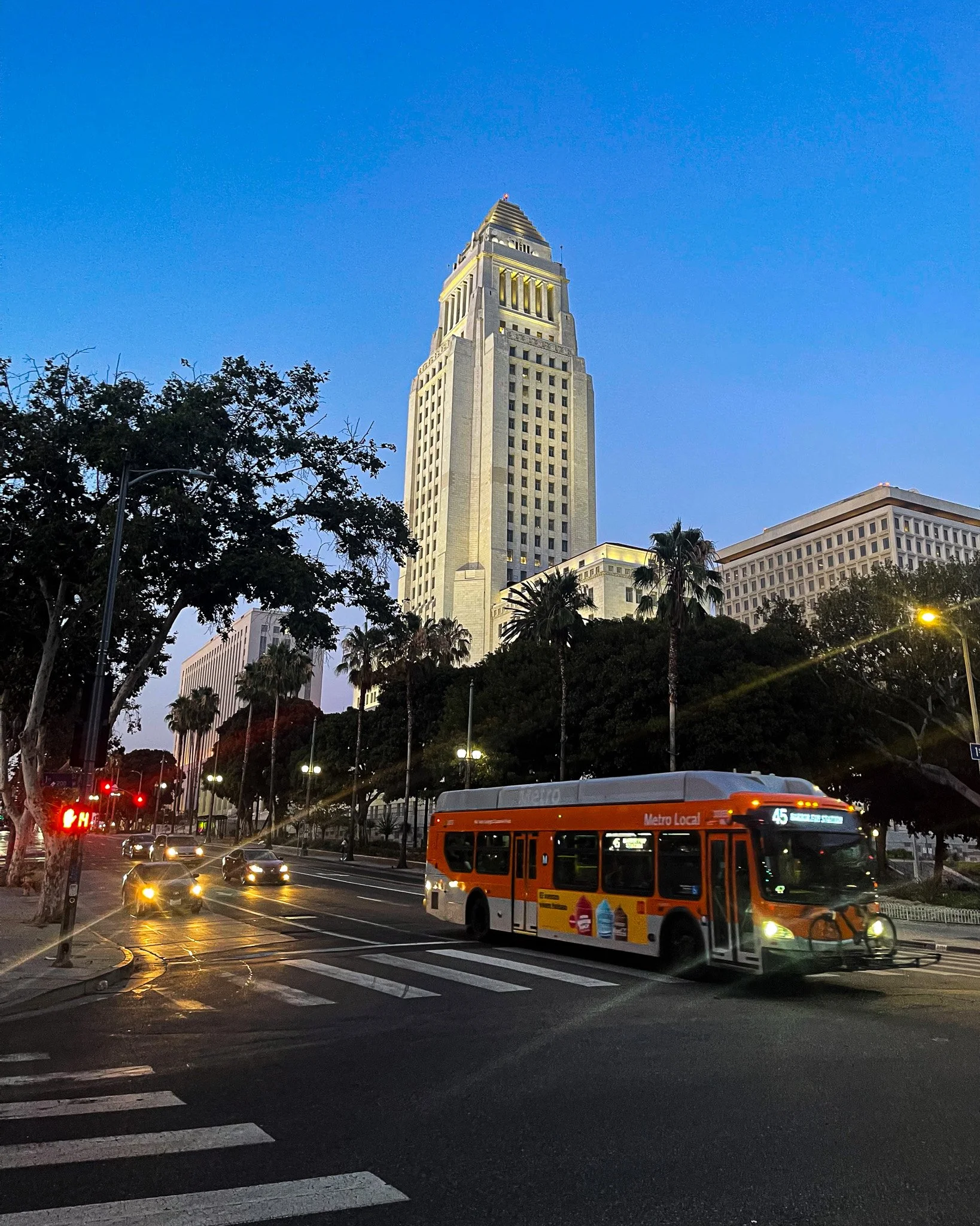 Orange Metro bus crossing a dusk intersection beneath the floodlit Los Angeles City Hall tower and street trees, 2023