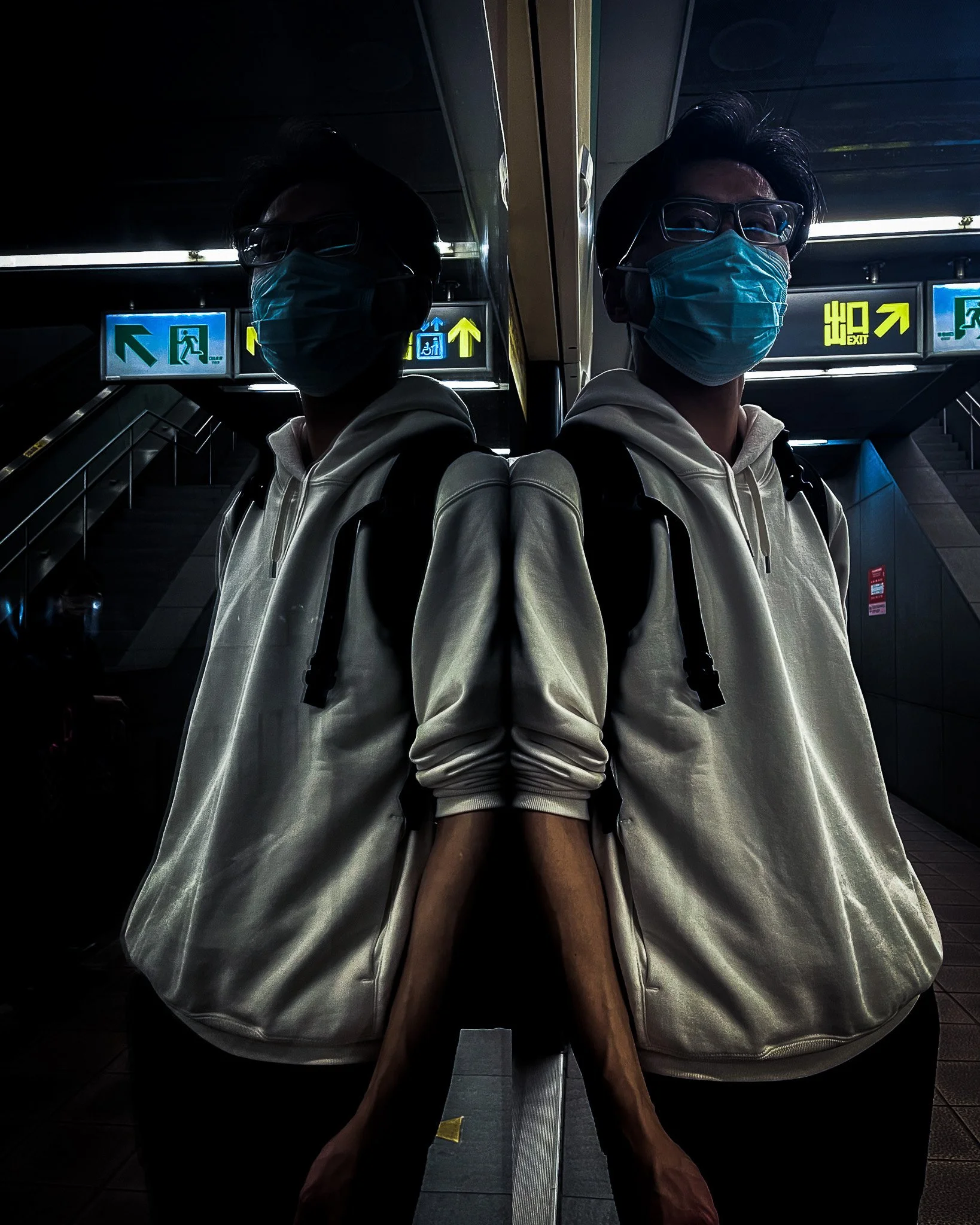 Person wearing a face mask, glasses, white hoodie, and backpack standing at an underground station, with their reflection in a mirror creating a symmetrical image.