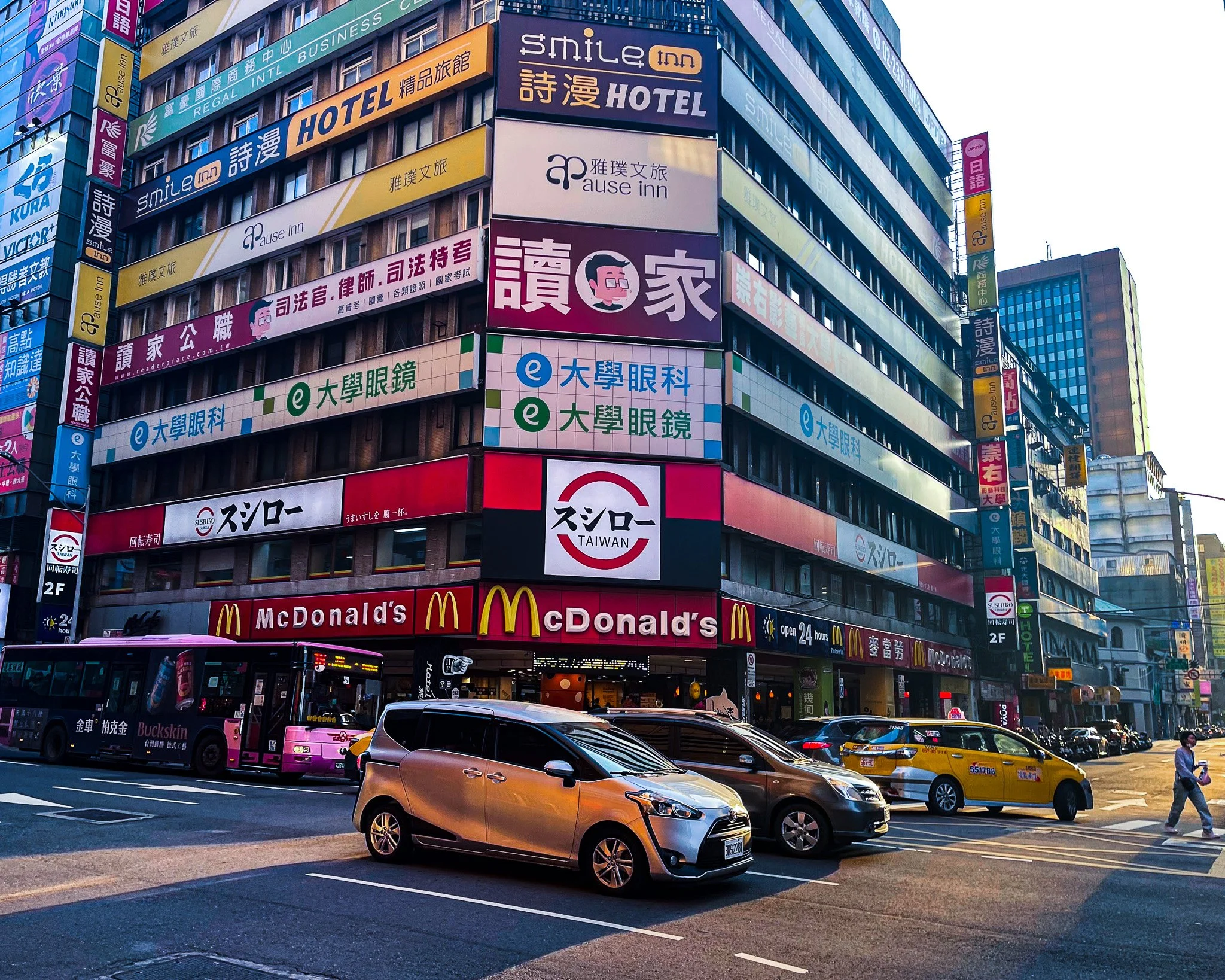 Urban street scene in Taipei with a purple bus, several parked and moving cars, a McDonald's restaurant, and numerous colorful signs on tall buildings advertising hotels, clinics, and other businesses.