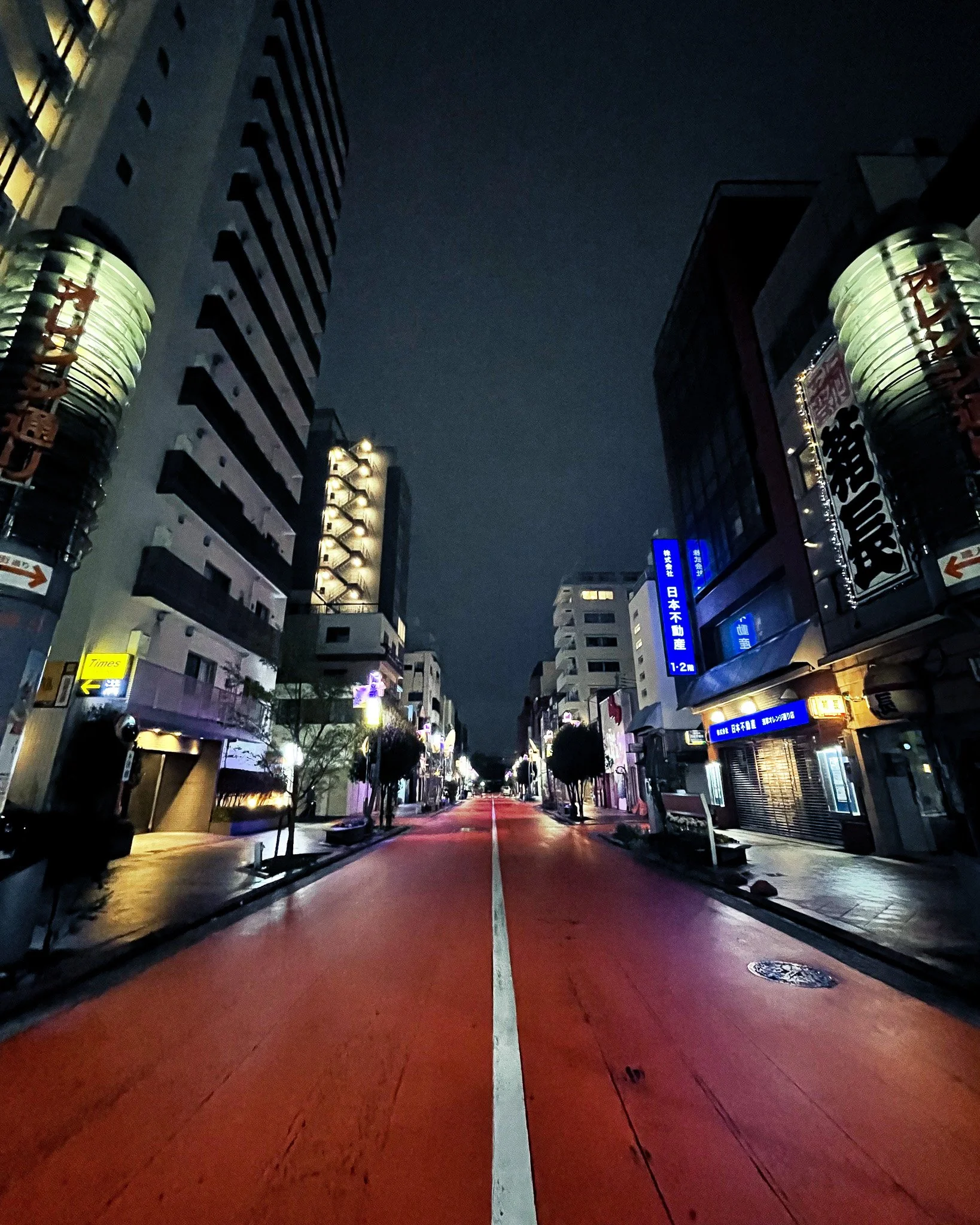 Empty city street at night with tall buildings, illuminated signs in Japanese, and a red road.