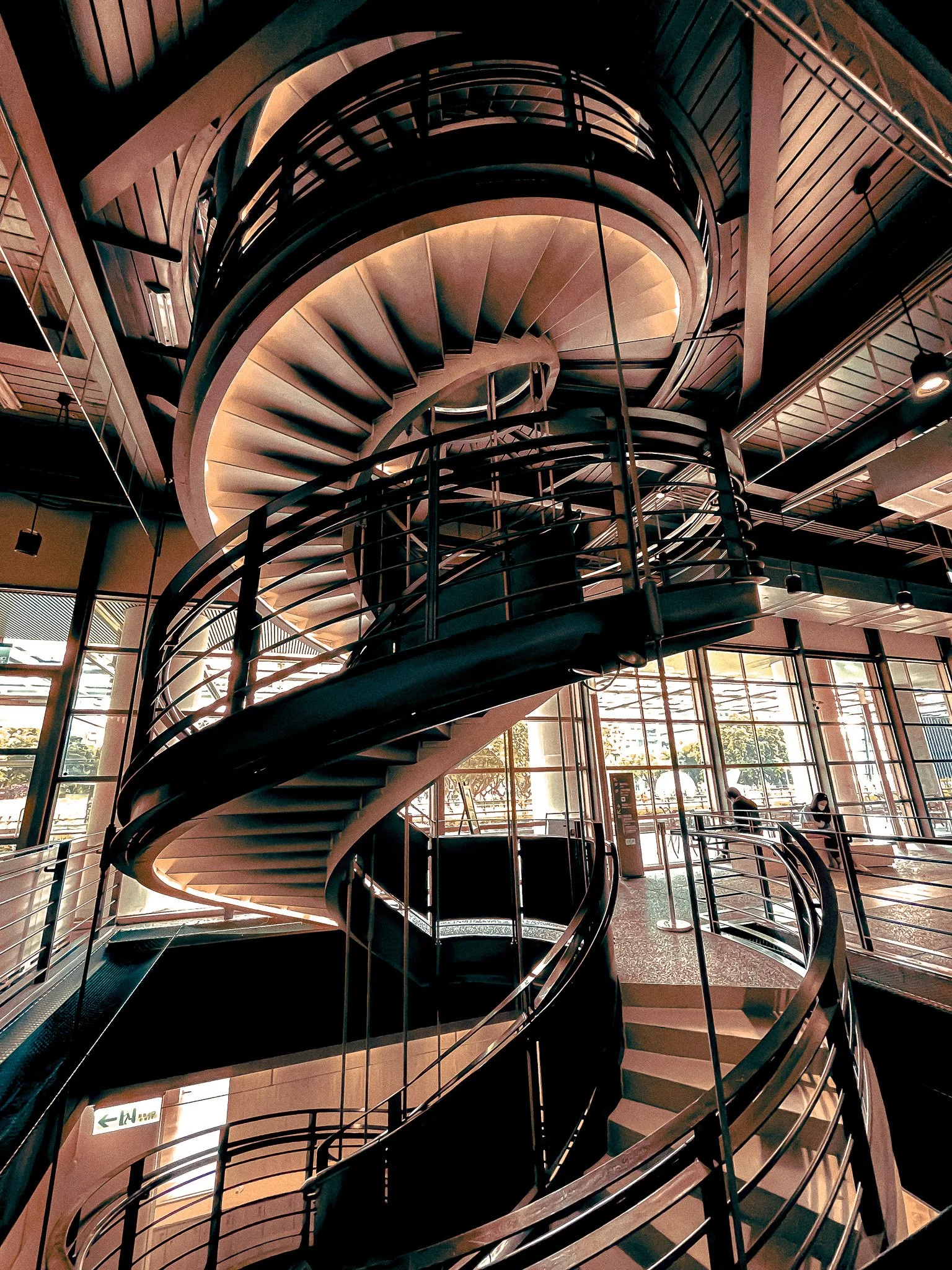 Modern spiral staircase inside a building with large glass windows and a wooden ceiling.
