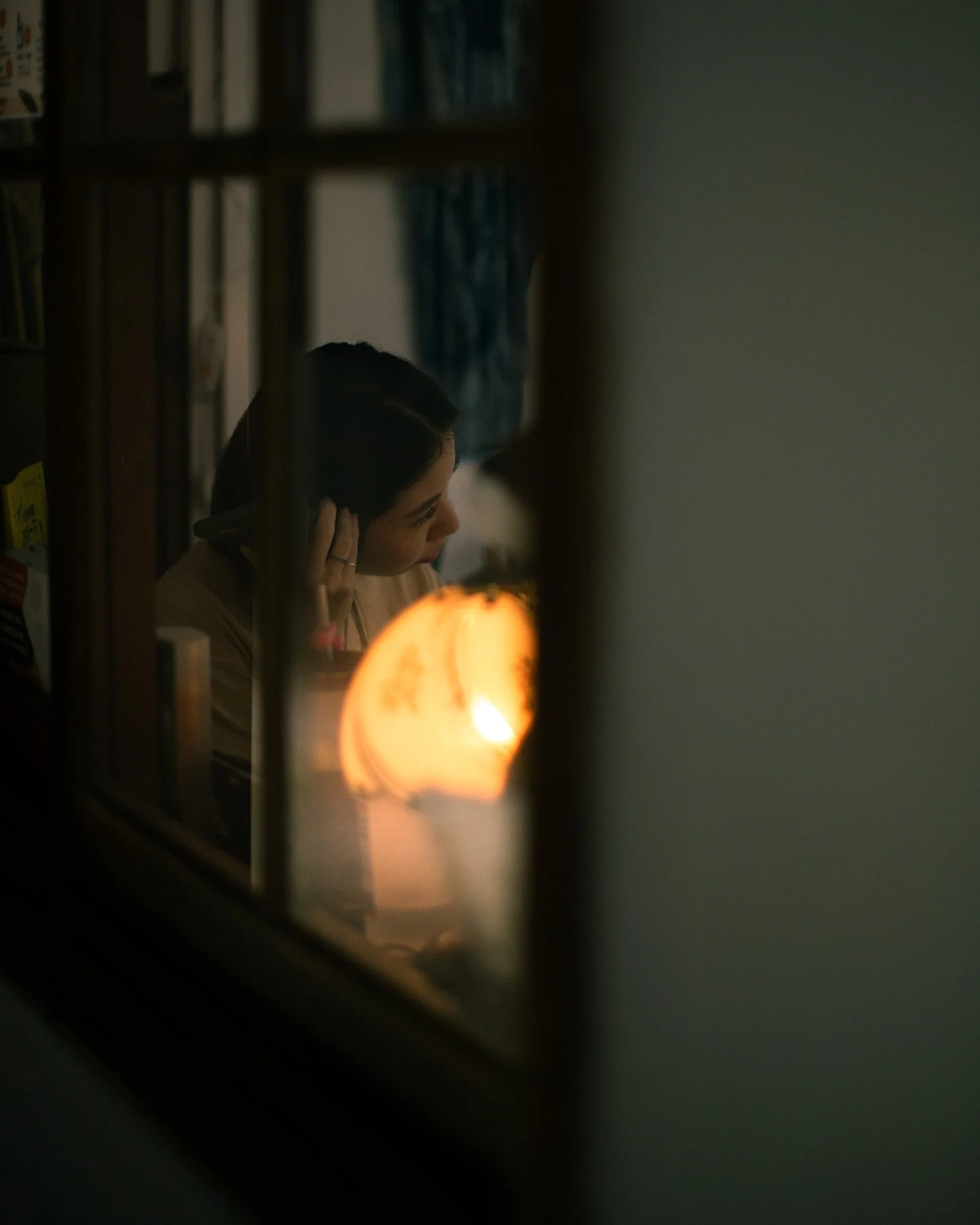 Person with dark hair seen through a grid, indoors with books and a warm light in the foreground