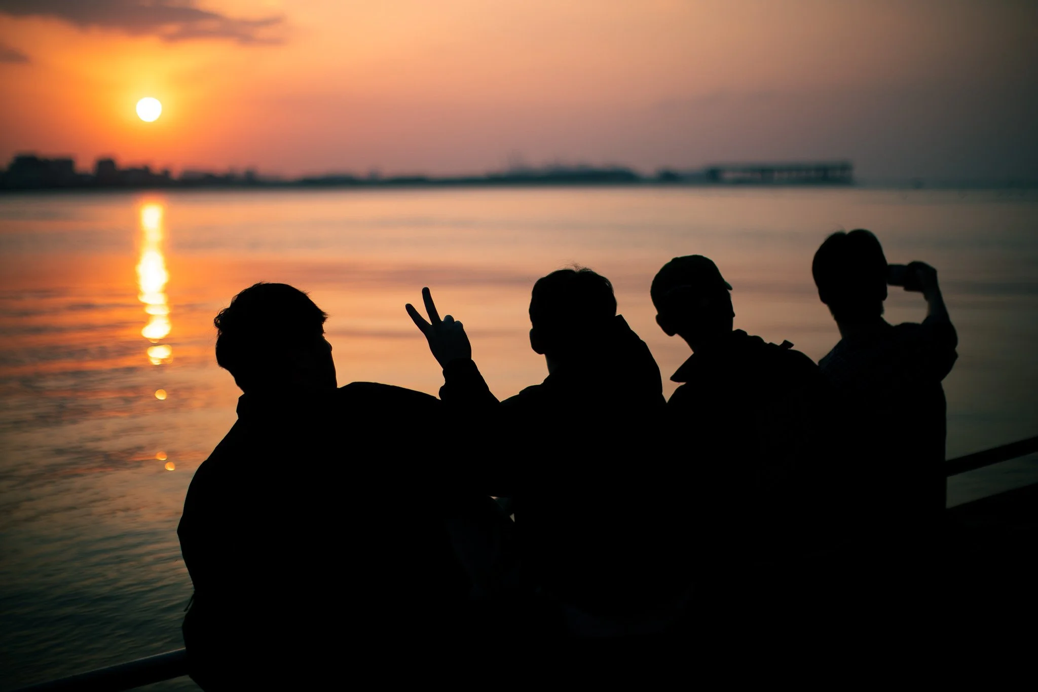 Four friends silhouetted against a sunset by the water, with one making a peace sign and another drinking from a bottle.