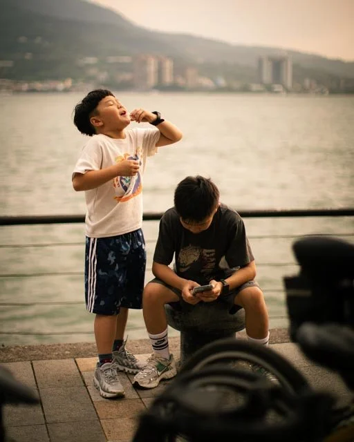 河畔欄杆旁的兩個男孩，一個站著吃點心，另一個坐著滑手機，台灣