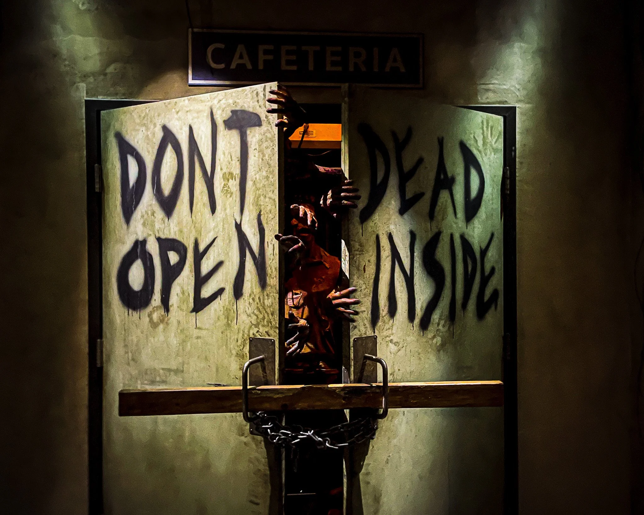a haunted house door with a sign above it that says 'CAFETERIA', and a creepy hand protruding through a broken section in the middle of the door, with the words 'DON'T OPEN DEAD INSIDE' painted on the door.