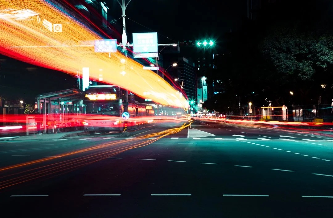 長曝光的車燈光軌劃過夜晚的路口，一輛公車停靠在站旁，台北