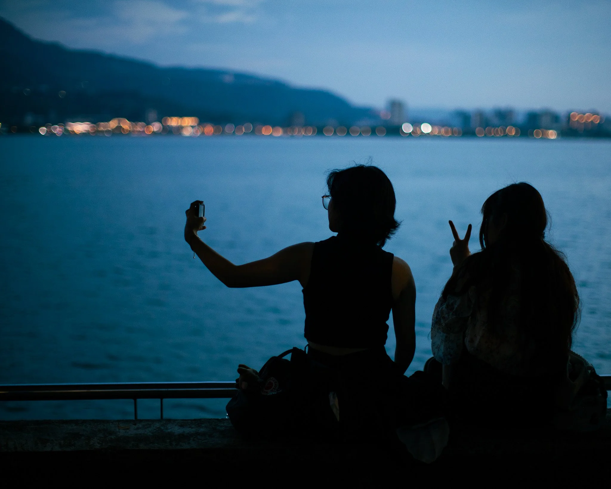 Two women sitting by the water at dusk, one taking a selfie and the other making a peace sign.