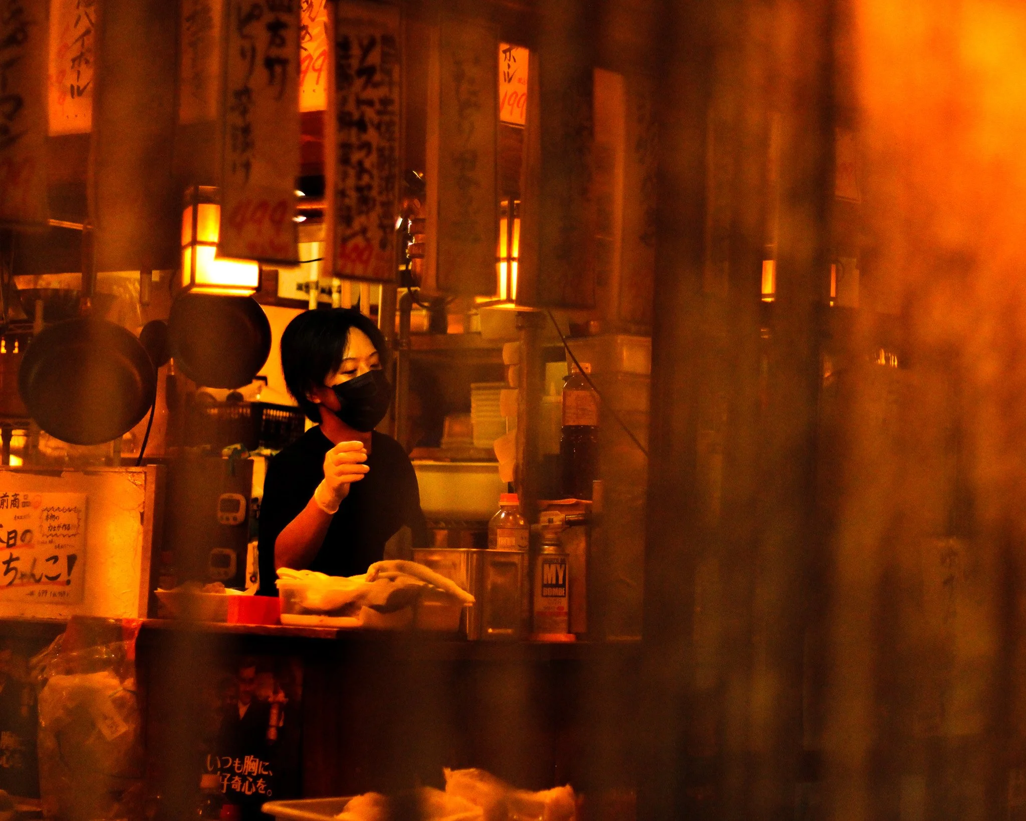 Person in a black face mask preparing food behind a counter in a warm, dimly lit eatery
