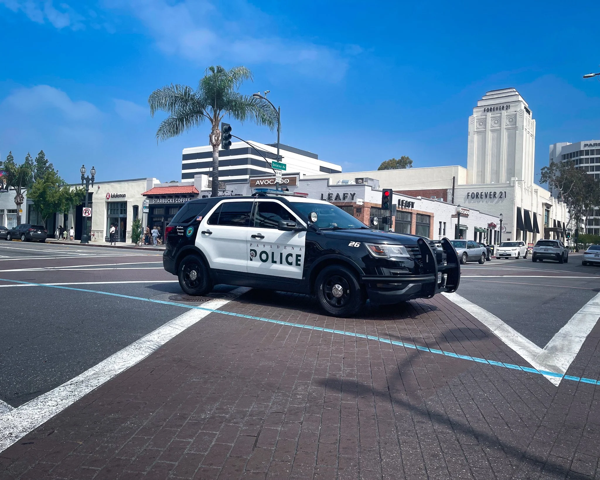 A black and white police car parked at a crosswalk on a city street. In the background are shops including Starbucks and Forever 21, tall palm trees, and a bright blue sky.