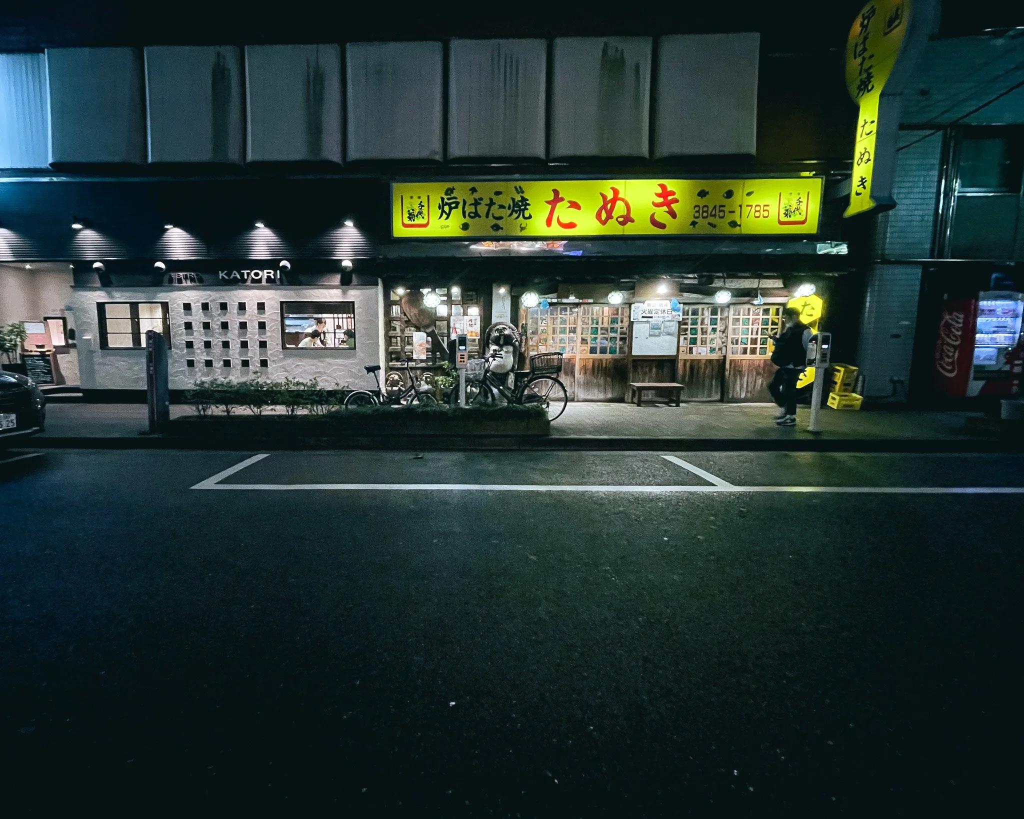 A Japanese restaurant storefront at night with yellow signage and bright interior lights, bicycles parked outside, and a person standing near a parking meter.