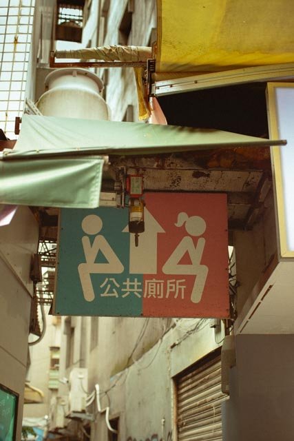 復古粉綠雙色的公共廁所指示牌懸在狹窄巷弄中，上方是褪色遮雨棚，台灣