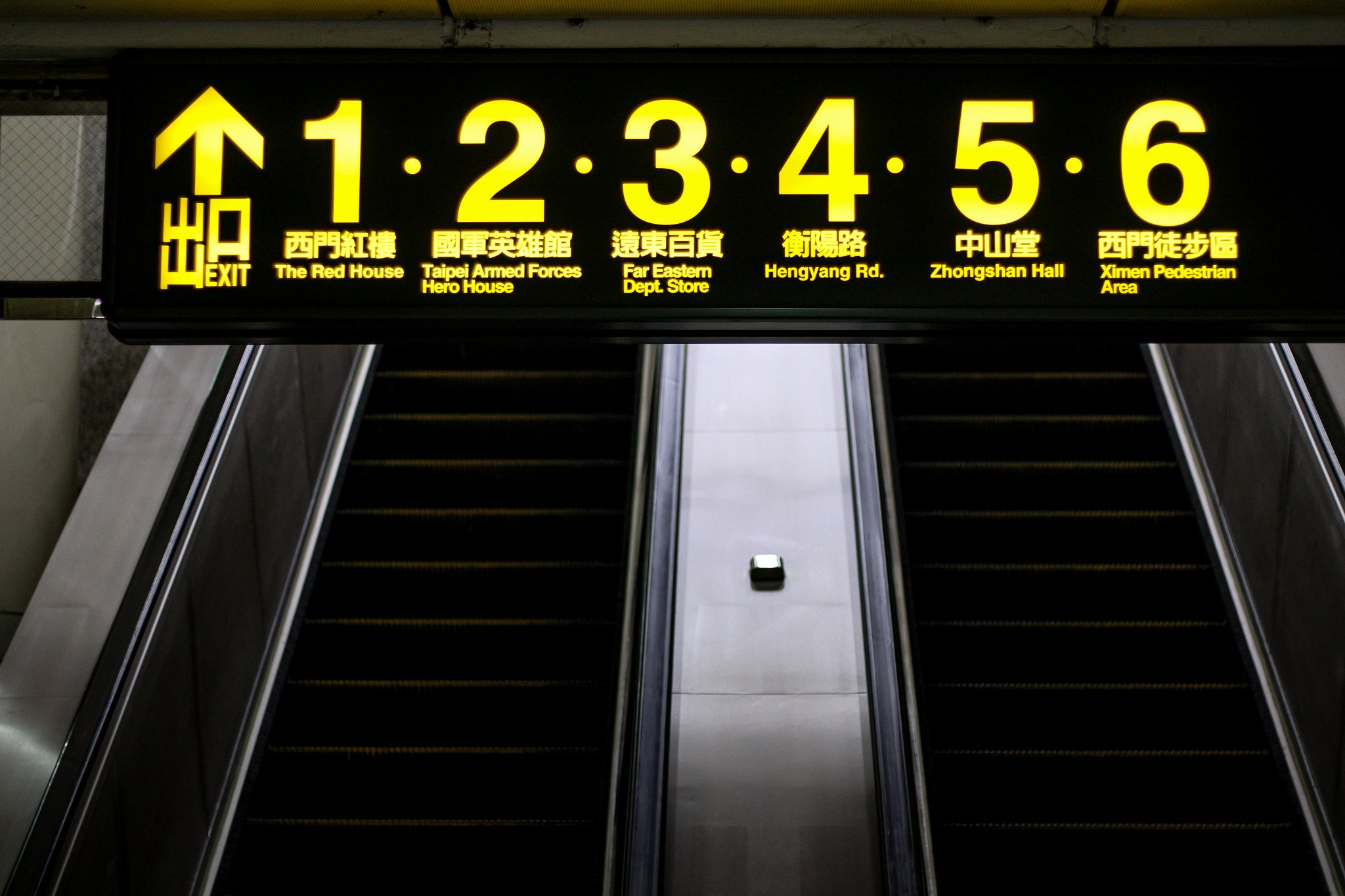 A signboard in a subway station shows directions to six different destinations, with each destination numbered from 1 to 6, along with their names in English and Chinese. Below the sign is an escalator leading downward.