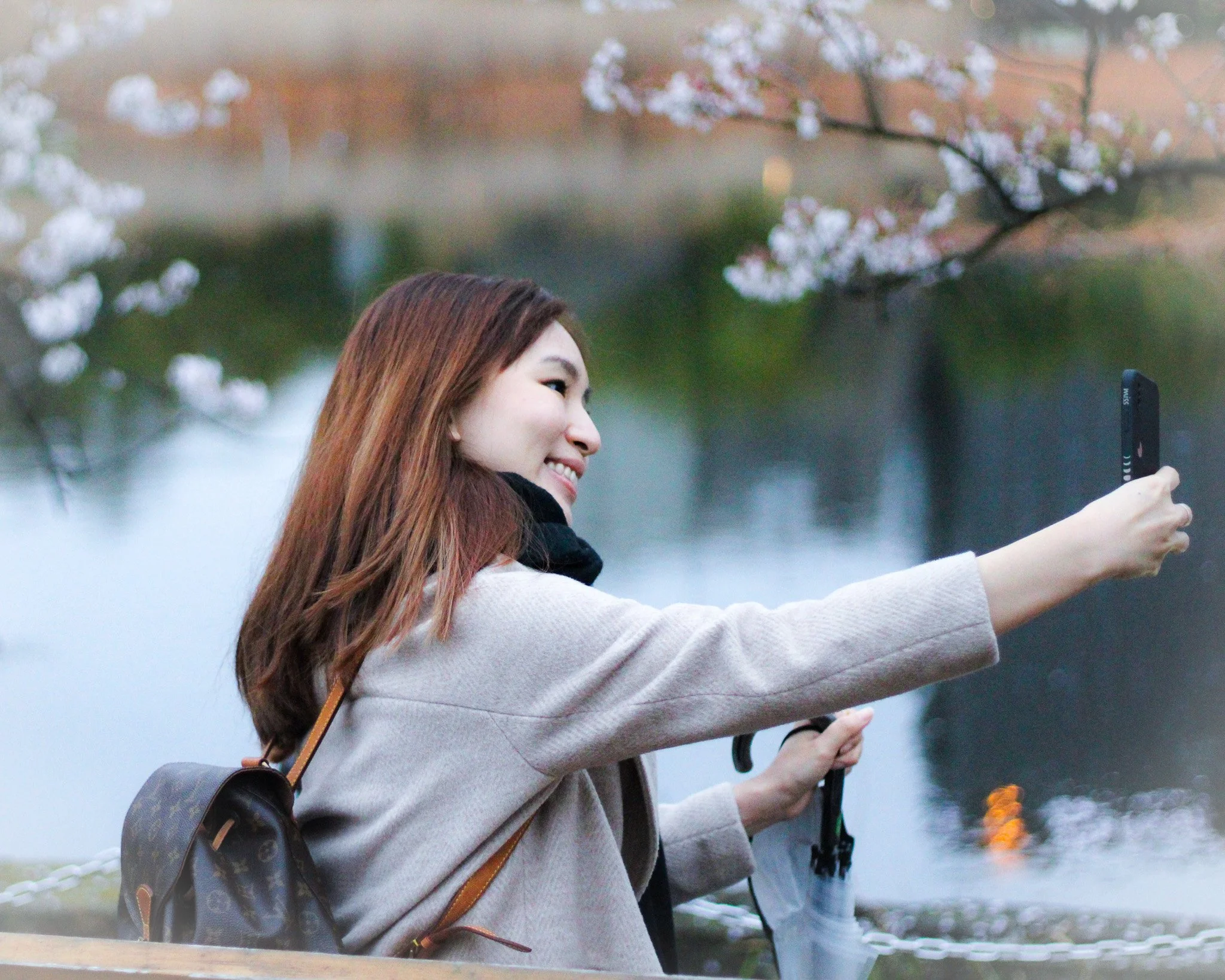 A woman with shoulder-length brown hair taking a selfie with her smartphone near a body of water.