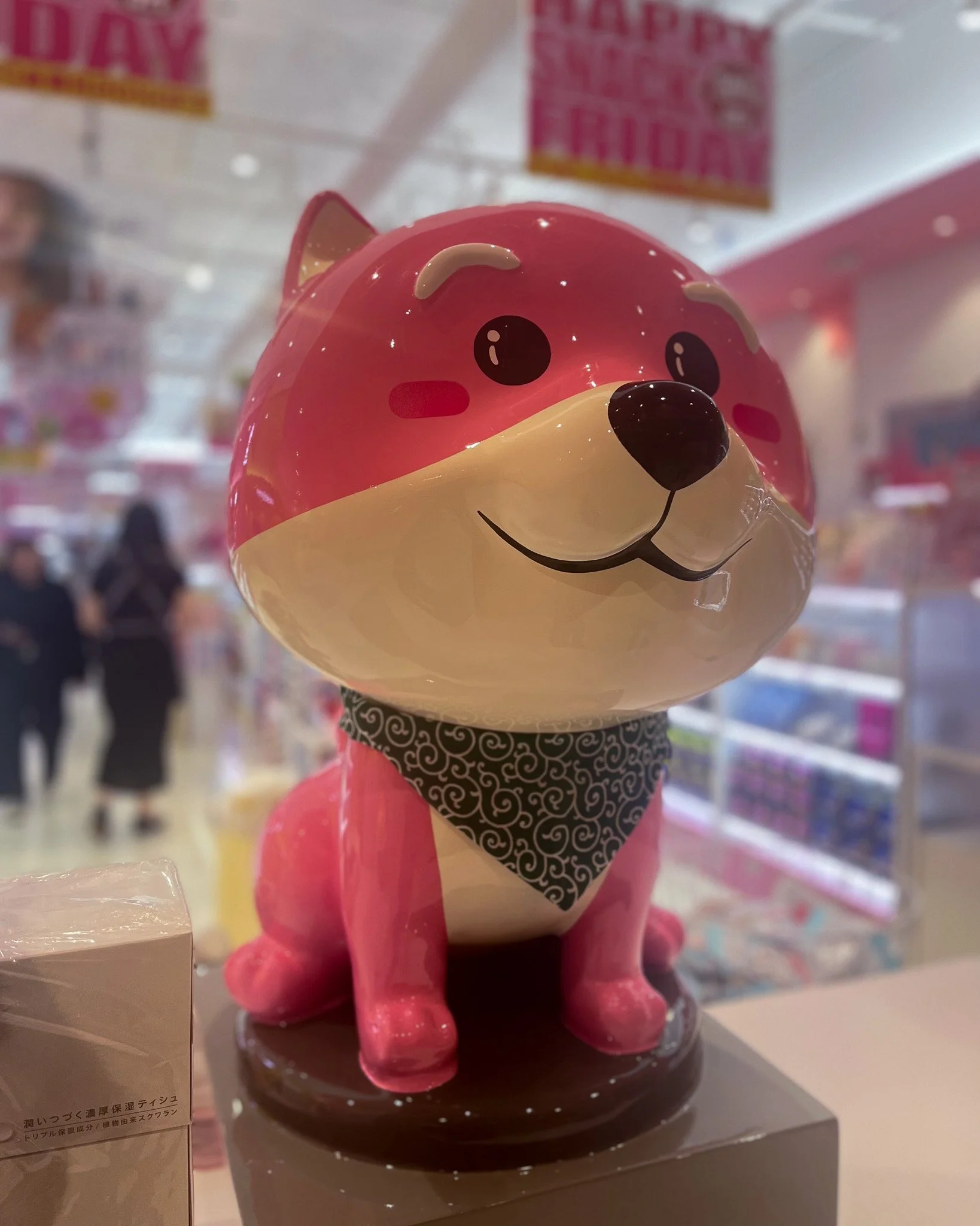 A pink and white dog-shaped figurine with cartoonish eyes, a black nose, and a smiling mouth, wearing a black patterned bandana, displayed on a shelf in a store.