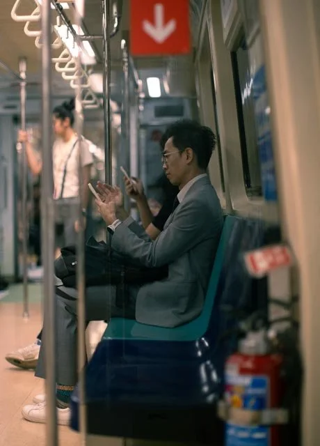 身穿灰色西裝、戴眼鏡的男子在捷運車廂裡低頭看手機，紅色箭頭標誌高掛，台灣