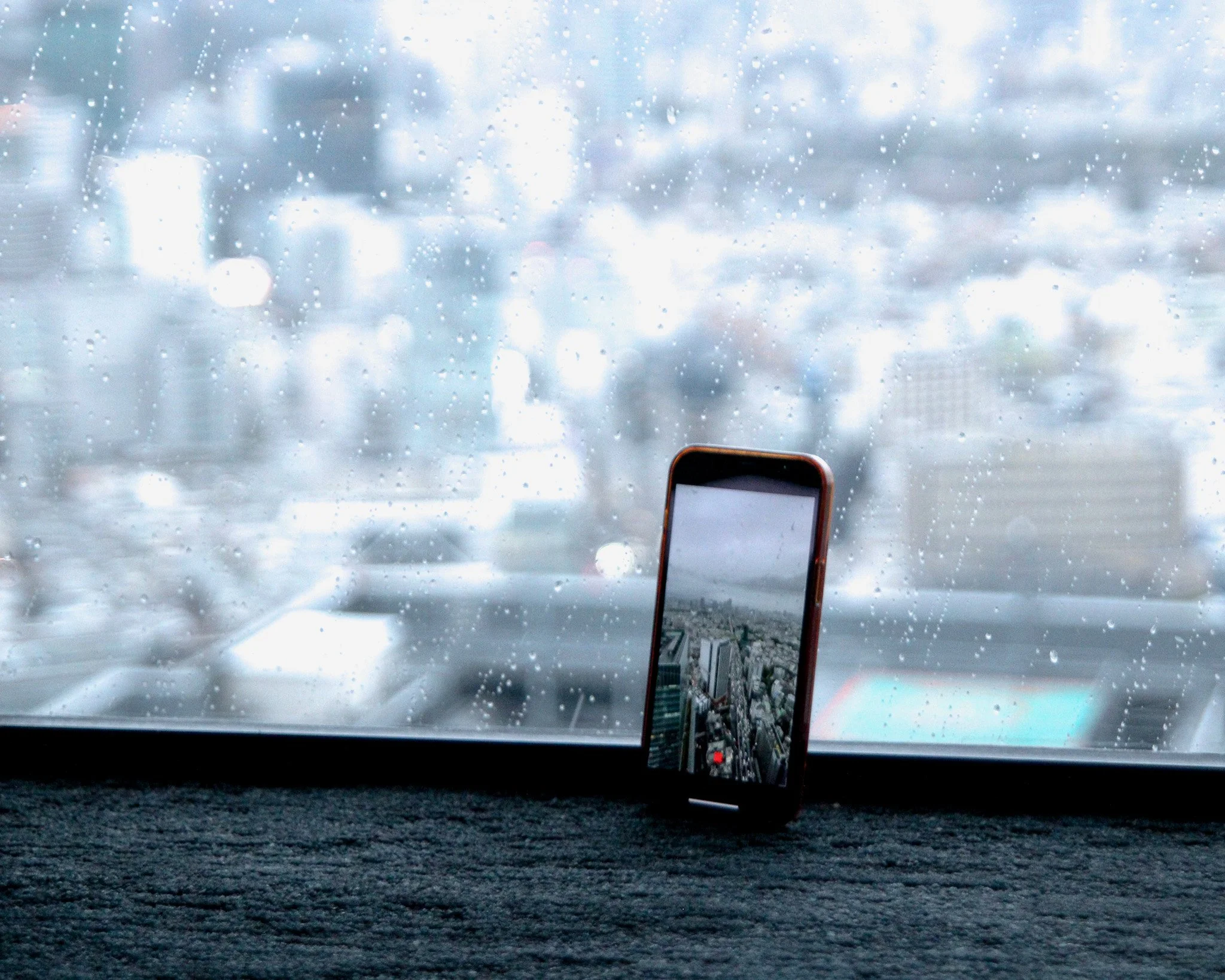 A smartphone with a cityscape wallpaper sitting on a black surface in front of a rain-covered window.