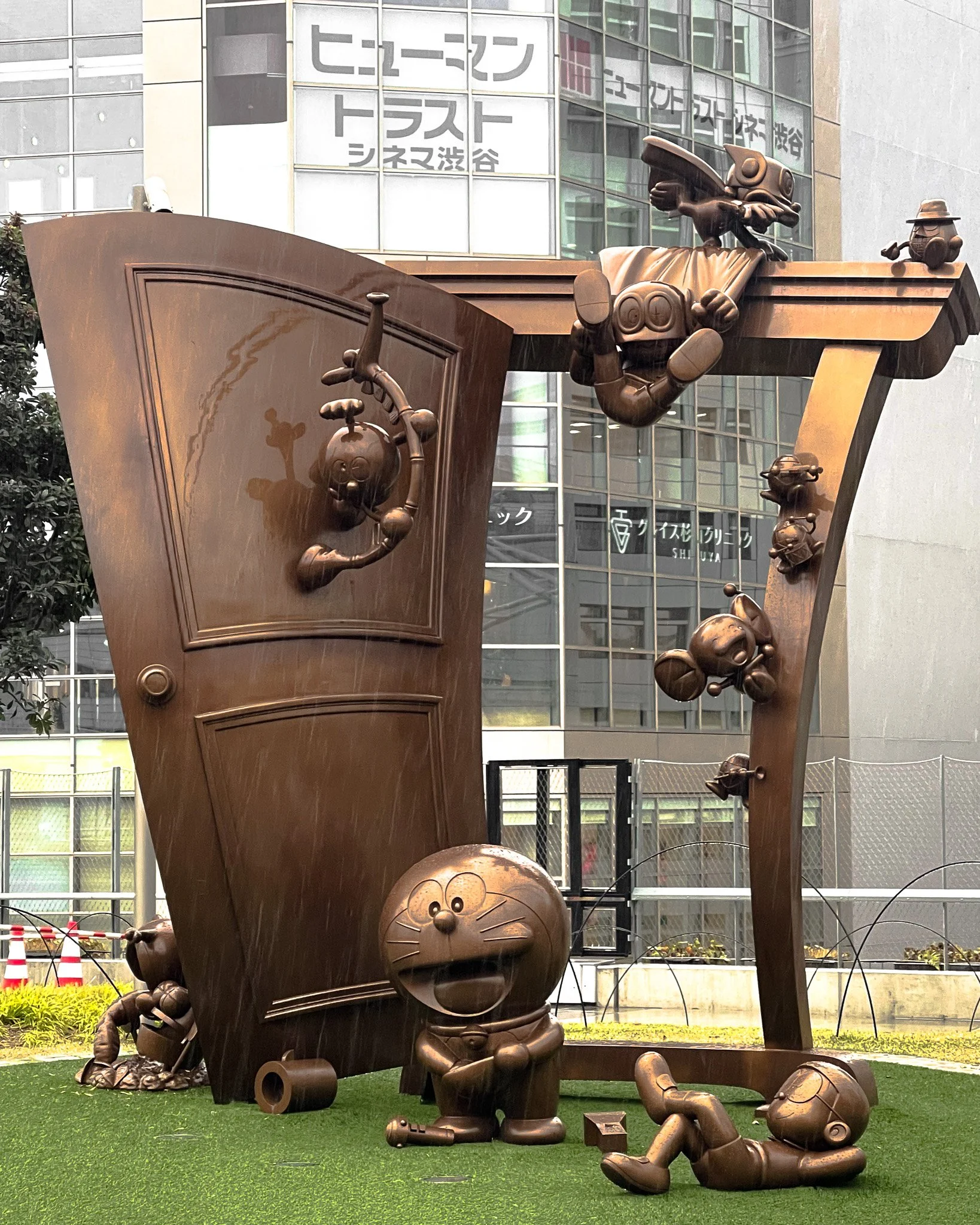Bronze statue of Doraemon and other characters from the Doraemon series around a large book, with some characters climbing and lying on it, set against an urban backdrop.