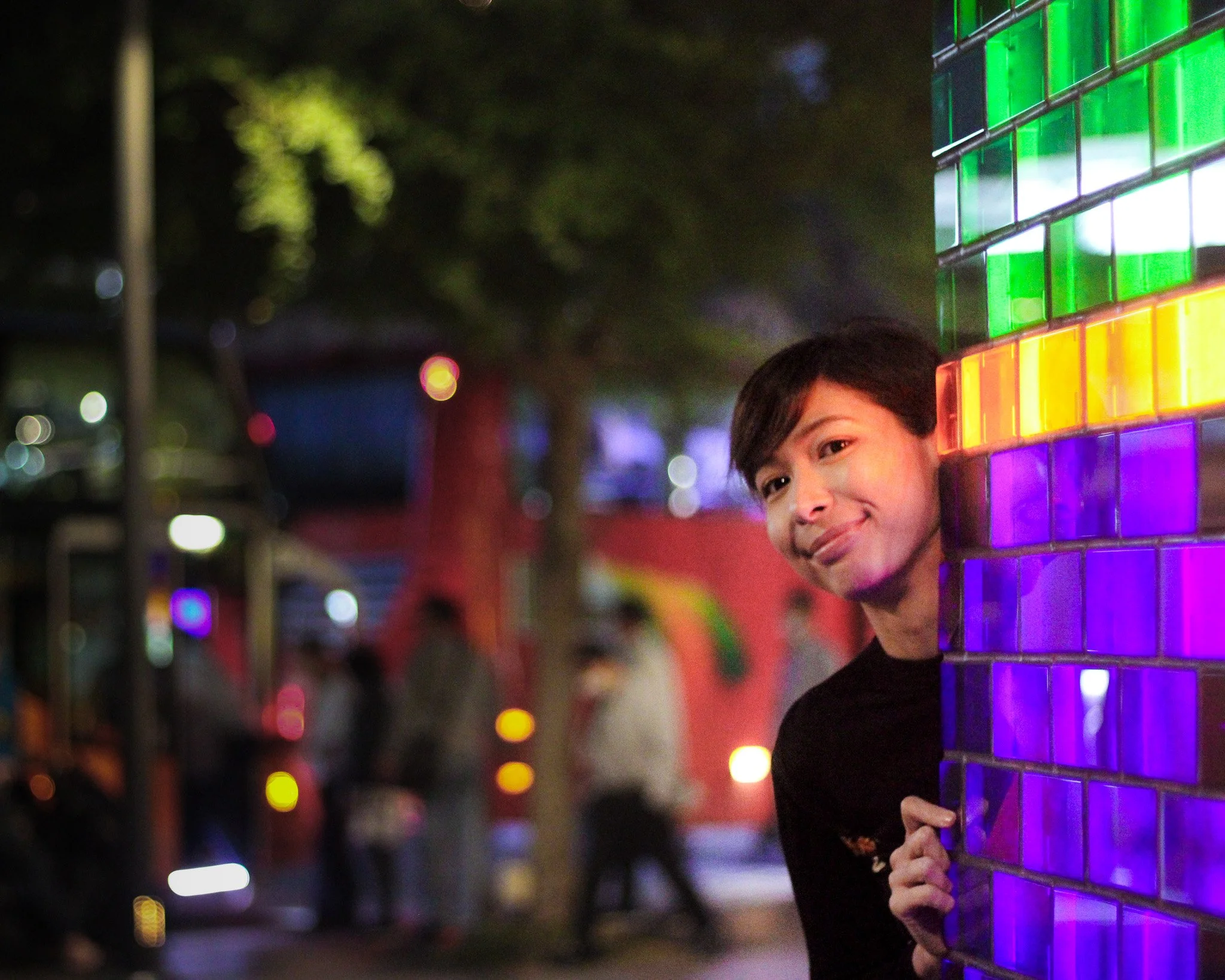 A person peeking from behind a colorful illuminated brick wall with rainbow-colored tiles.