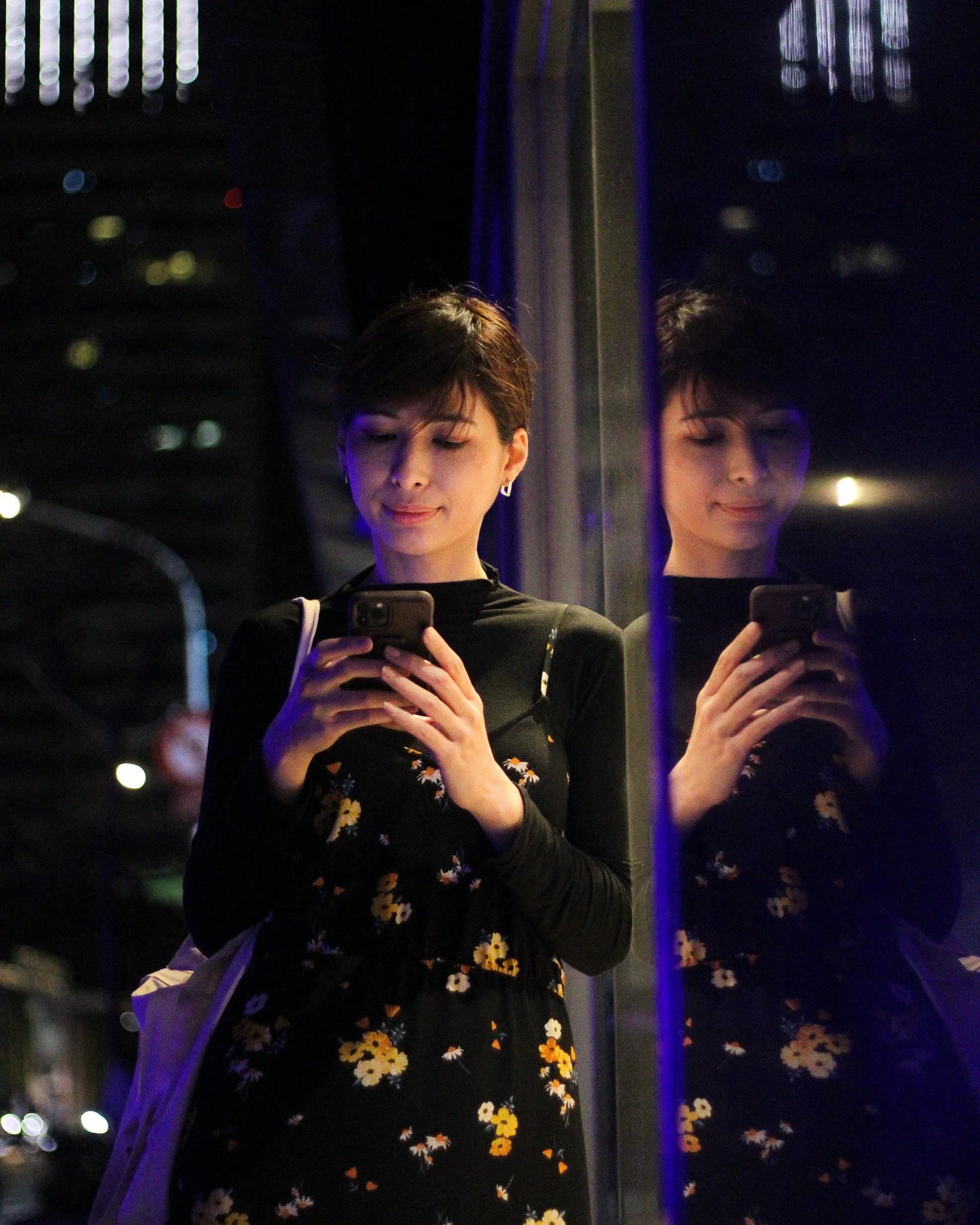 Young woman taking a selfie with her phone, standing in front of a mirror at night, wearing a black floral dress, with city lights in the background.