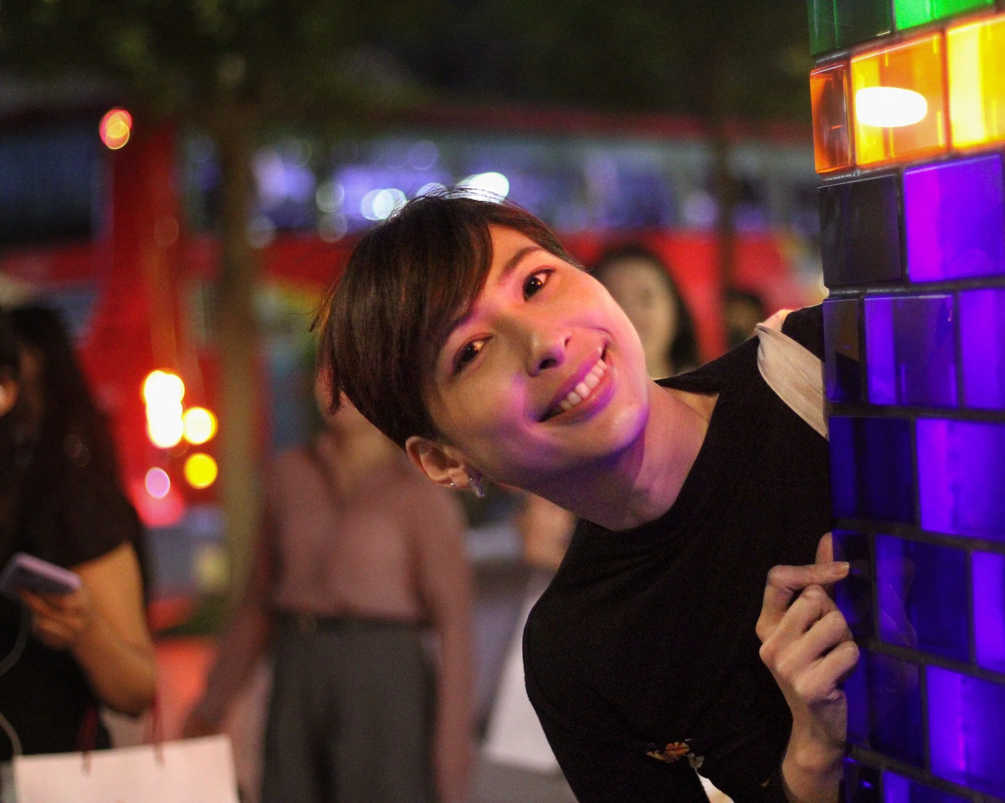 A smiling person with short brown hair peeks out from behind a colorful illuminated panel at an evening outdoor event, with other people and blurred lights in the background.