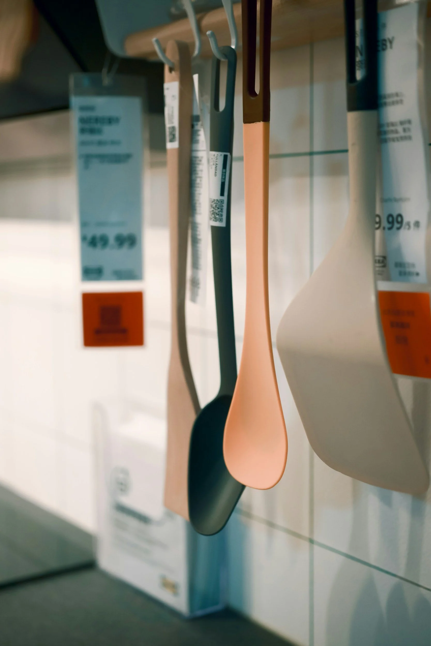 Close-up view of kitchen utensils hanging on a rack, including a black spoon, a peach-colored spoon, and a black spatula, with price tags and a store shelf in the background.