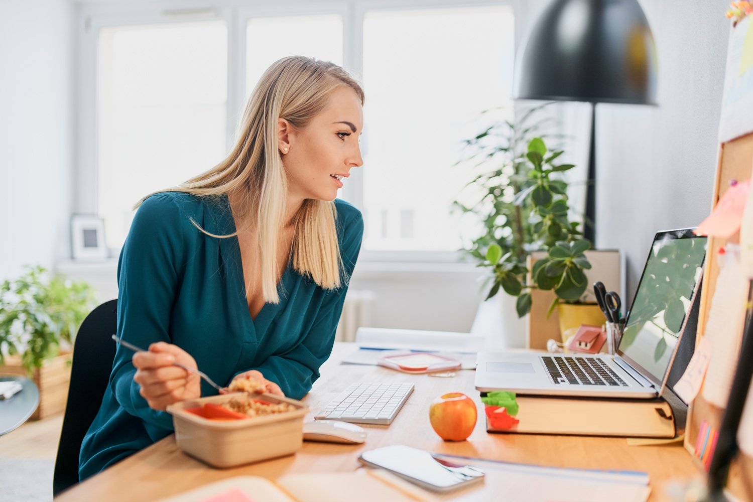 Woman working at a desk with a laptop, apple, and food container, in a bright, modern office.