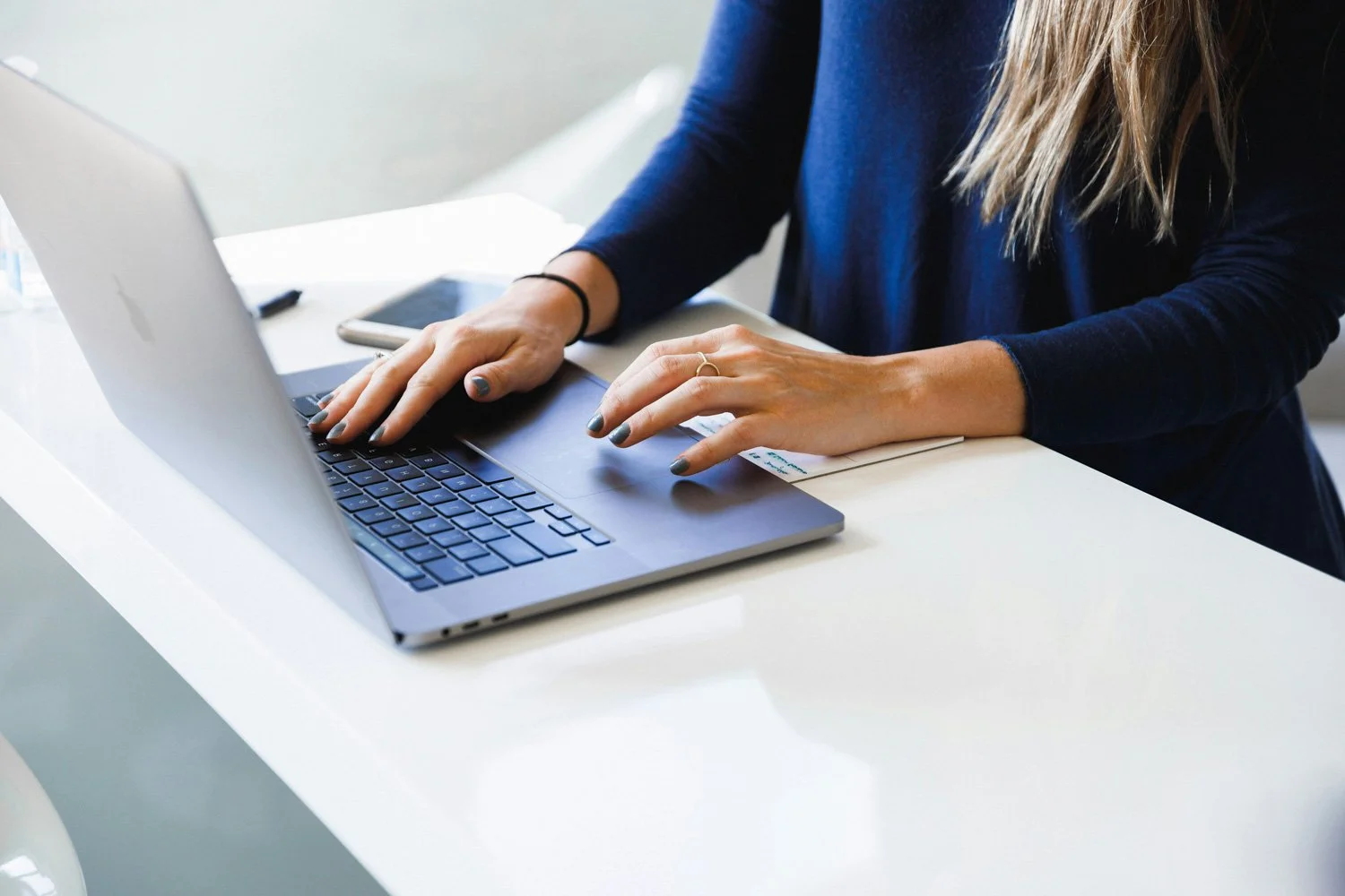 Person with long blonde hair typing on a laptop at a white desk, surrounded by a smartphone and notes.