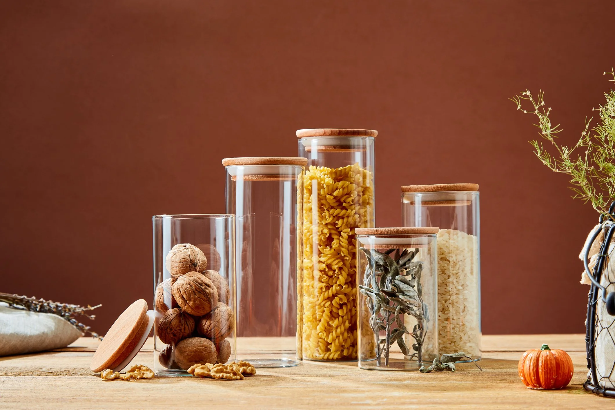 Several glass jars with wooden lids containing pasta, walnuts, sage, and rice are arranged on a wooden table with a brown background. A small pumpkin and some plant sprigs are also visible.