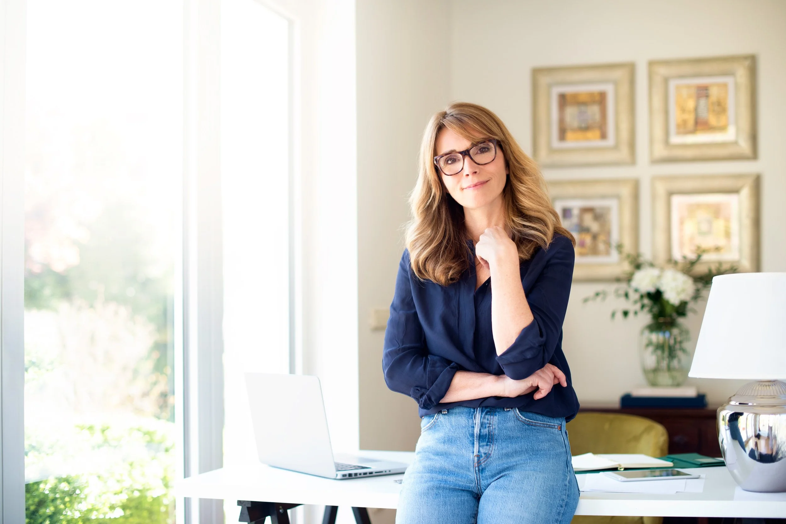 A woman in glasses dressed in a dark blue shirt and light jeans, standing in a bright room next to a desk with a laptop, a bread maker, and some art on the wall behind her.