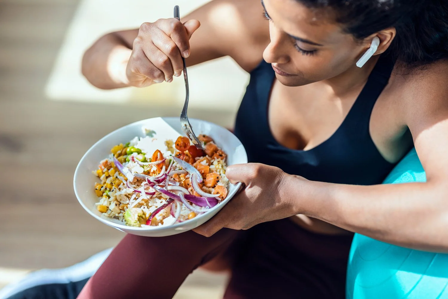 A woman with earbuds is sitting and holding a plate of mixed vegetables and rice while eating with a fork.