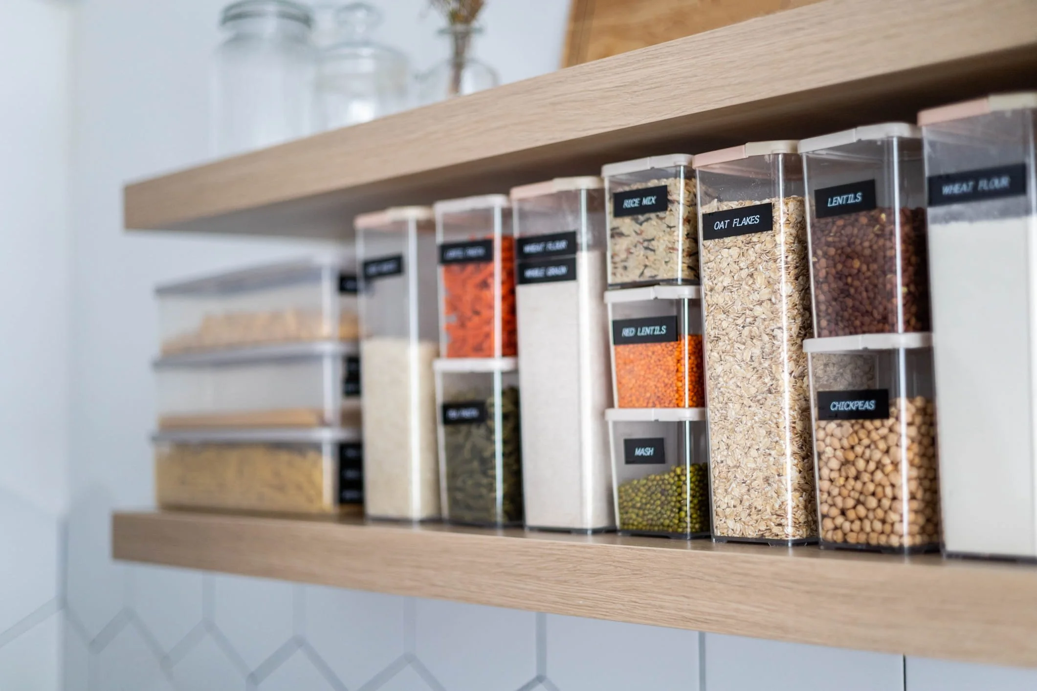 Bottle containers filled with dry grocery items like rice, lentils, oatmeal flakes, chickpeas, and wheat flour are arranged on a wooden shelf in a pantry or kitchen.