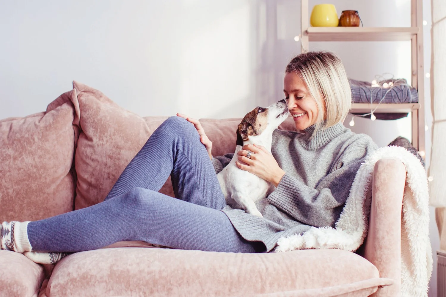A woman sitting on a pink couch cuddling a small dog, smiling at each other, in a cozy living room with a shelving unit and string lights in the background.