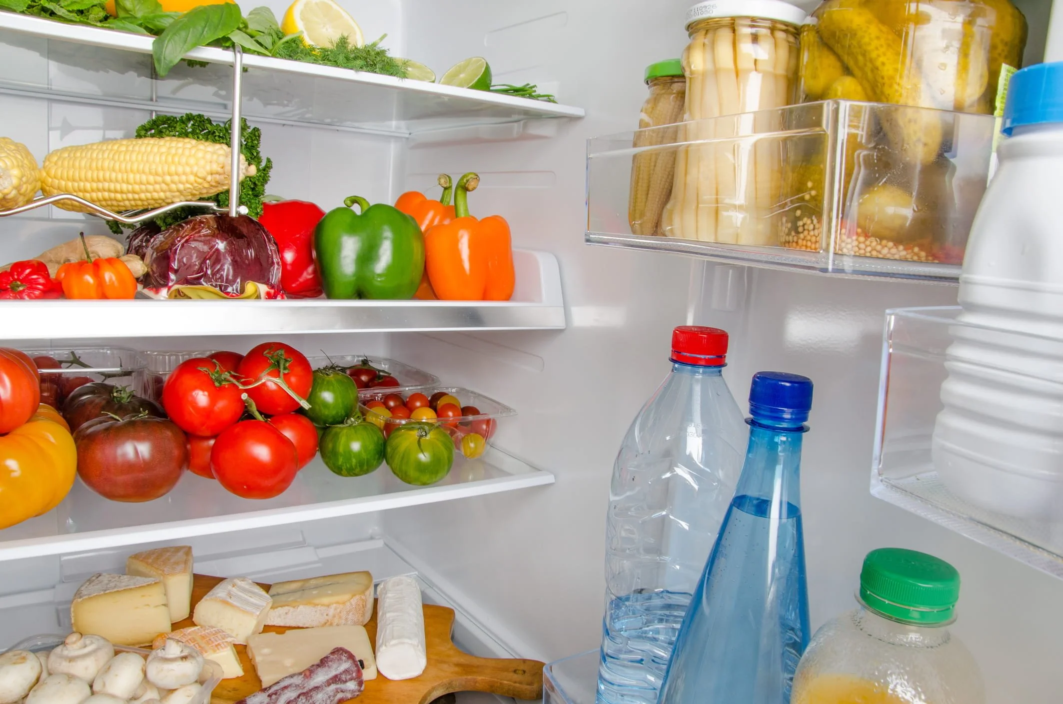 Inside of a refrigerator with fresh vegetables on the shelves, including corn, green and orange bell peppers, tomatoes, and red and yellow peppers, as well as jars of pickles and bottled water.