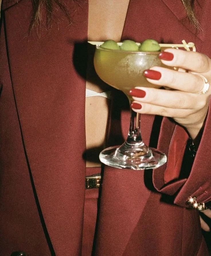 Person holding a cocktail glass with green olives, wearing red nail polish and a burgundy blazer.