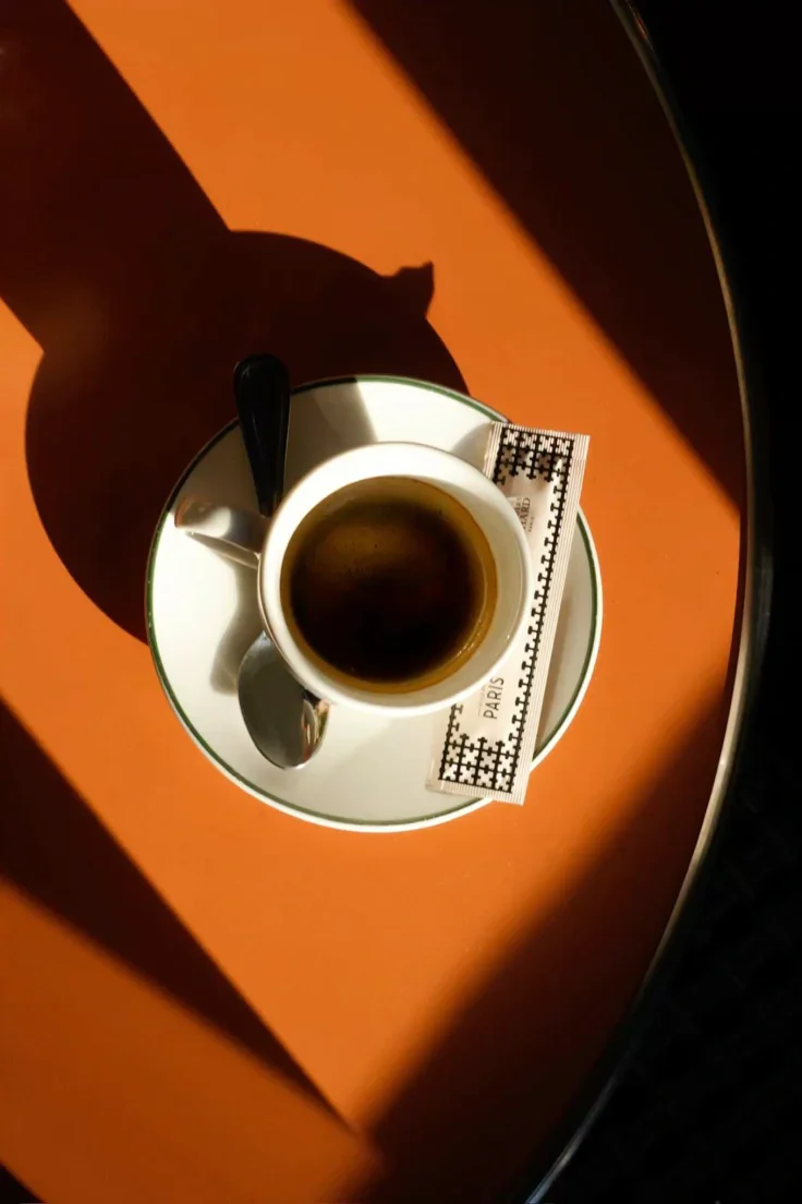 A cup of black coffee on a saucer with a spoon and a small packet of sugar, placed on an orange table with a shadow.