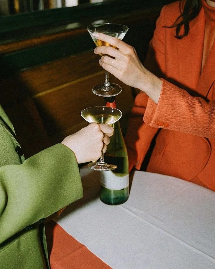 Two people in colorful suits clinking wine glasses with a green bottle on a table, indoors.