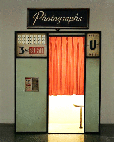 Photo booth with orange curtain, framed sign reading 'Photographs', and signs advertising 3 photos for $1.50 and 'While-U-Wait' service.